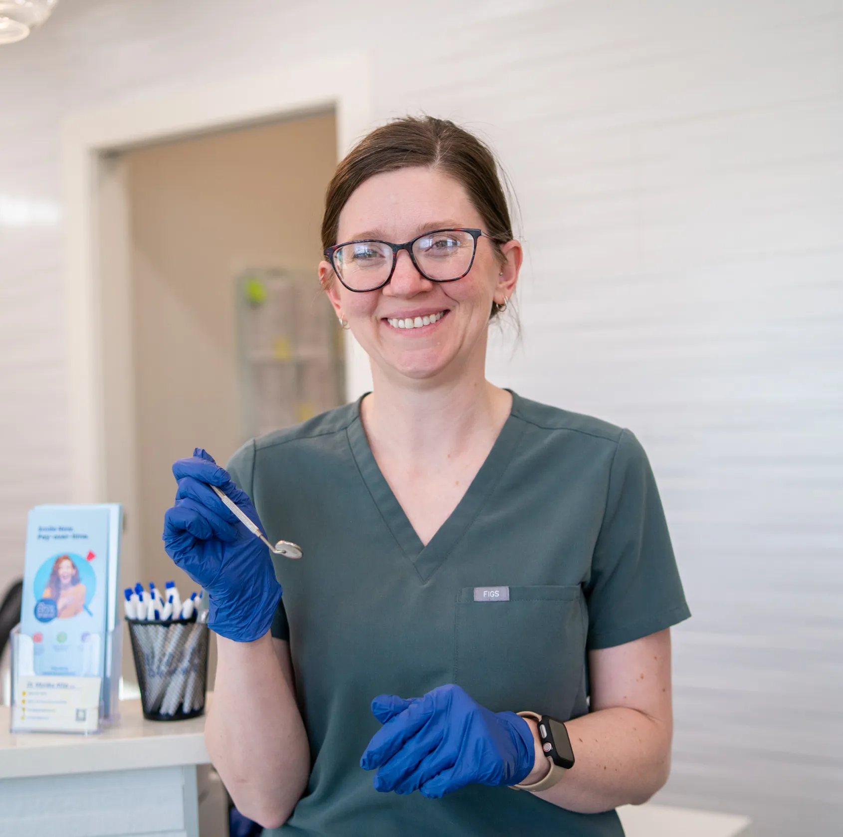 Smiling female dental professional in green scrubs and blue gloves holding a dental mirror in a dental clinic.