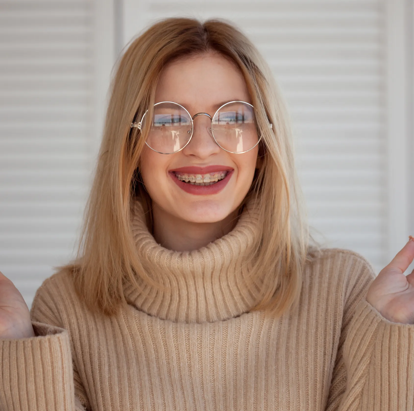 Smiling young woman with blonde hair, wearing round glasses and metal braces, dressed in a beige turtleneck sweater.