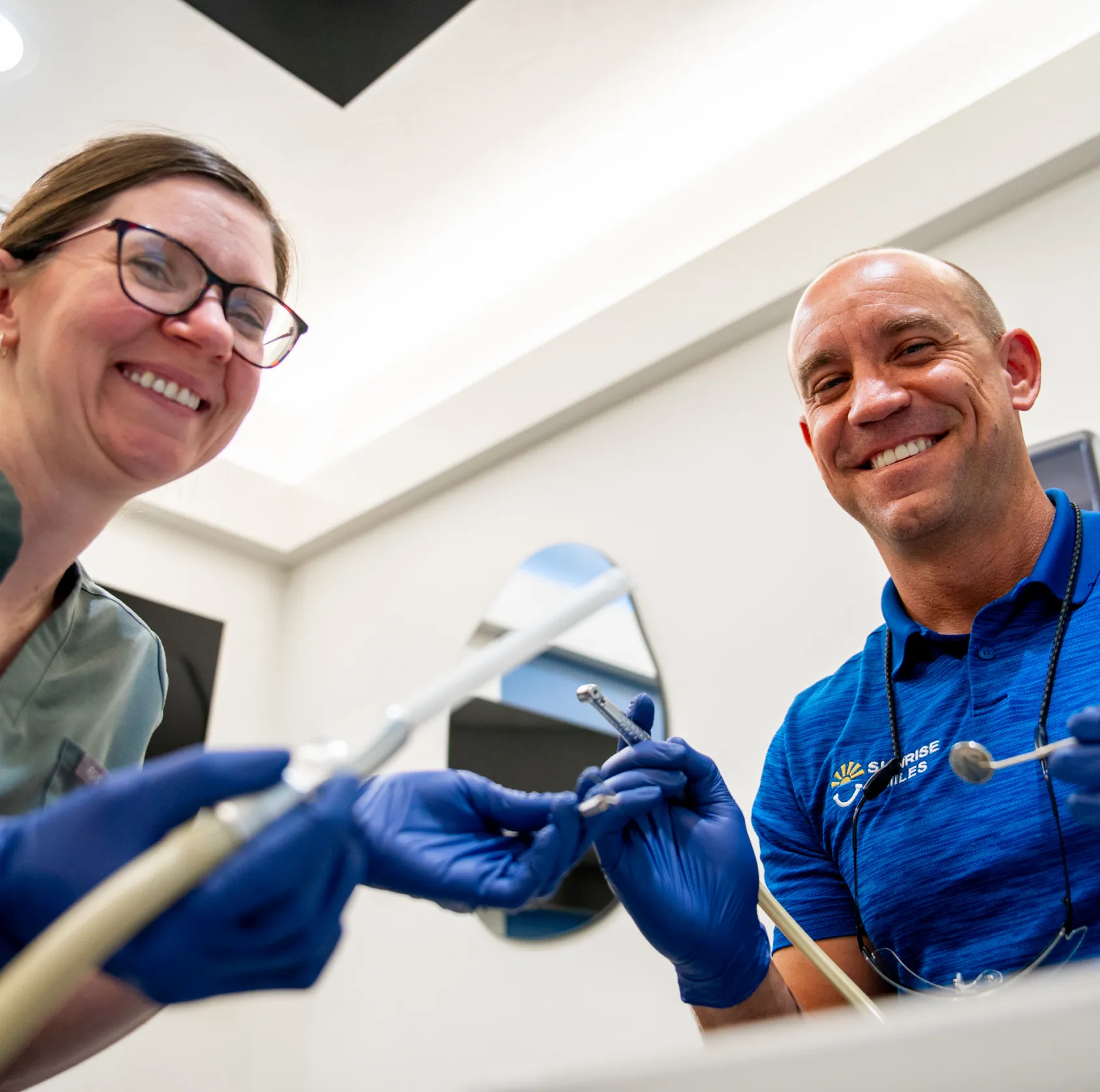 Two smiling dental professionals wearing blue gloves holding dental tools in a clinic.