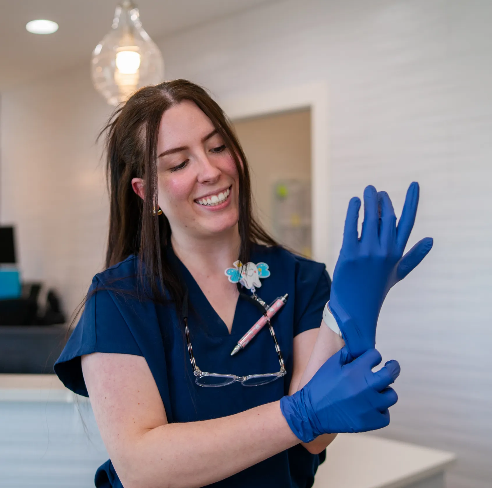 Smiling healthcare worker in navy scrubs putting on blue gloves in a clinical setting.