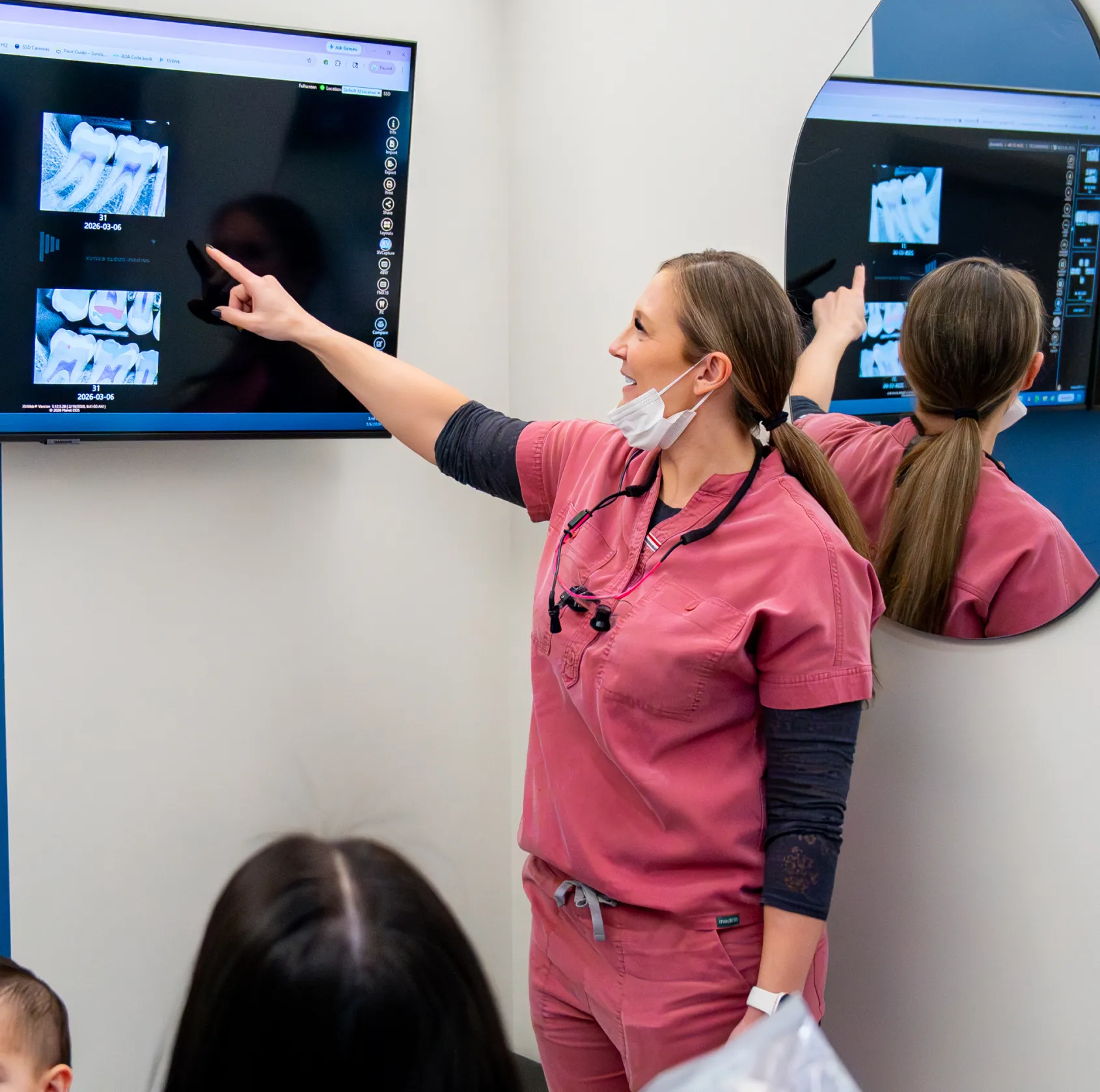 Female dentist in pink scrubs pointing at dental X-rays displayed on a wall-mounted screen during a patient consultation.