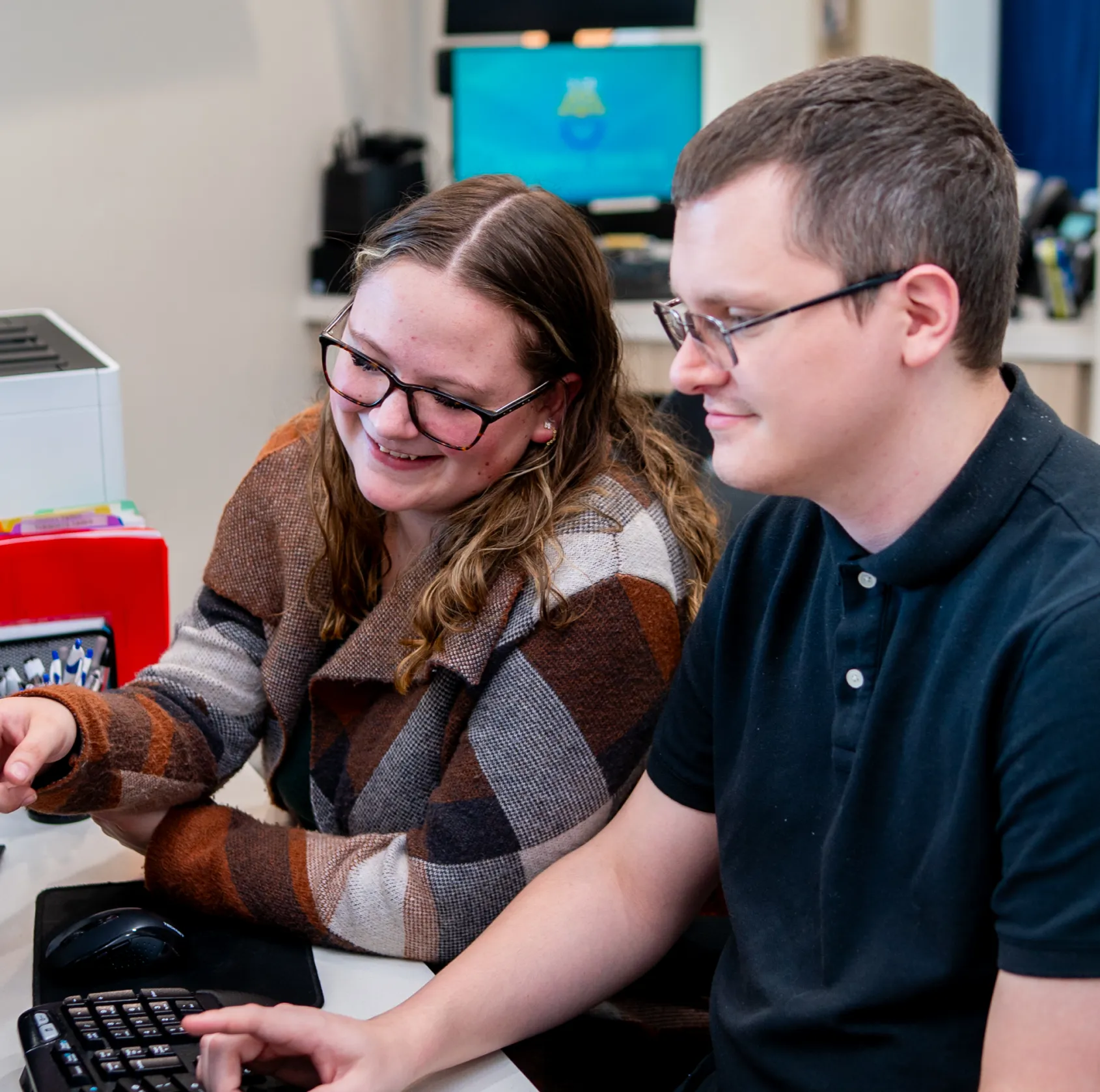 Two people sitting at a desk working on a computer together, one pointing at the screen and both smiling.
