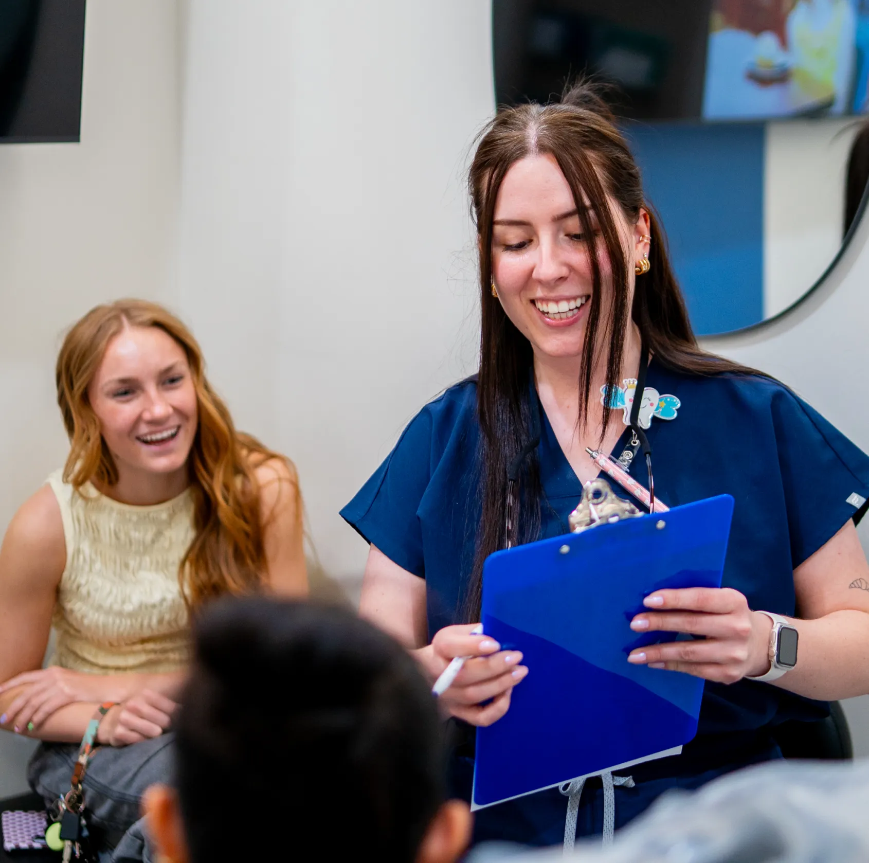 Smiling healthcare professional in blue scrubs holding a clipboard, talking to patients in a clinic setting.