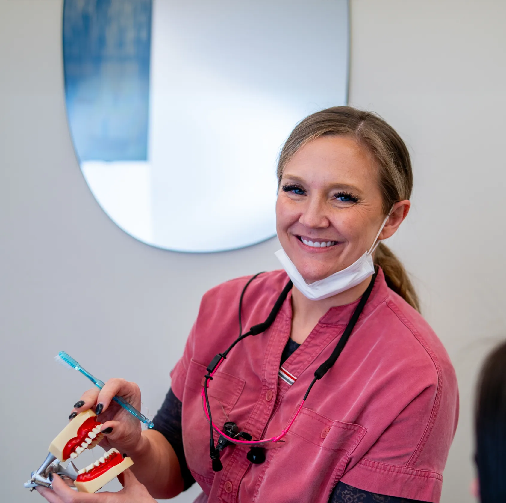 Smiling female dentist in pink scrubs holding a dental model and toothbrush, demonstrating oral care.