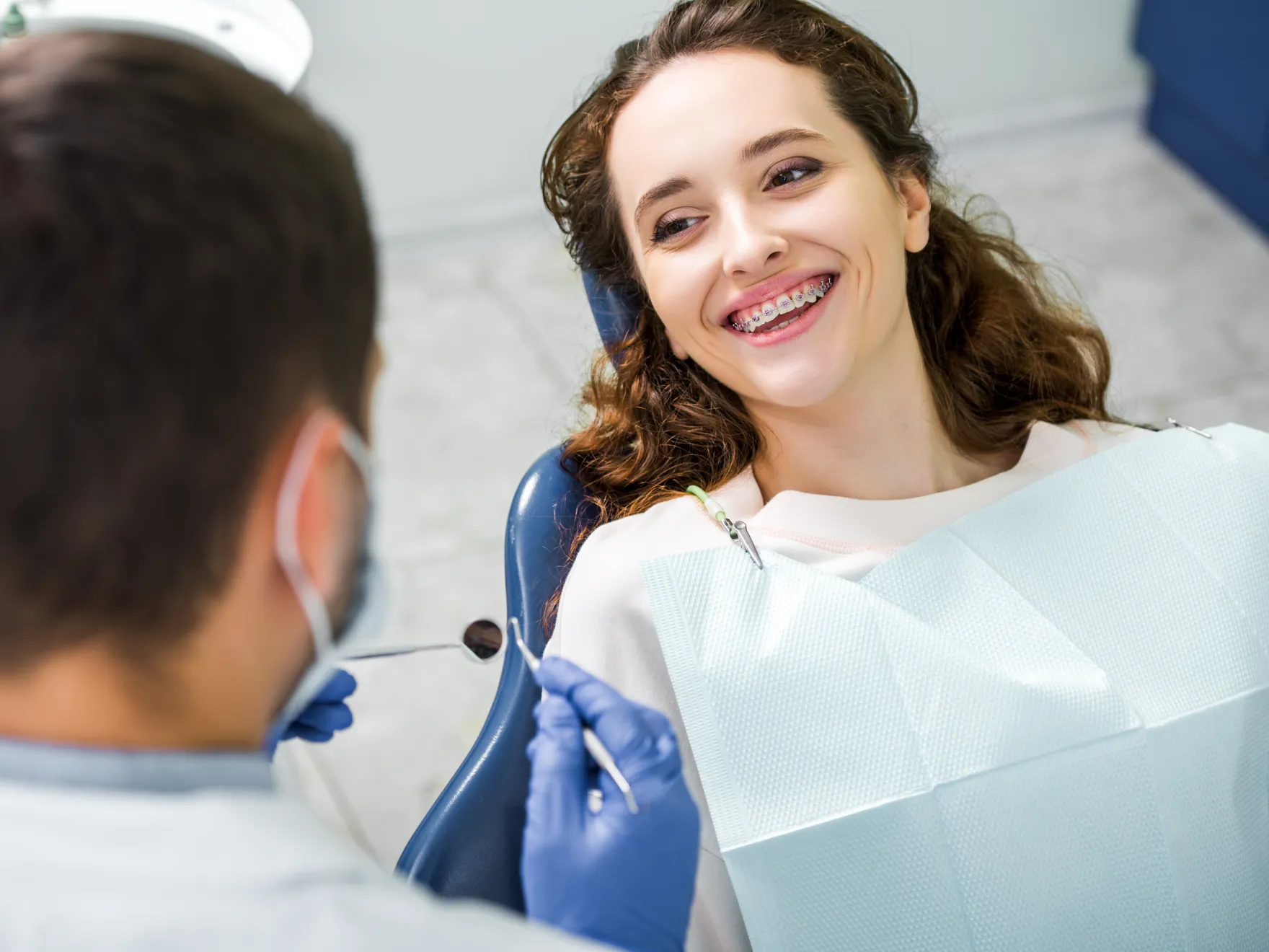 Woman with metal braces smiling while sitting in a dental chair with a dentist examining her teeth.