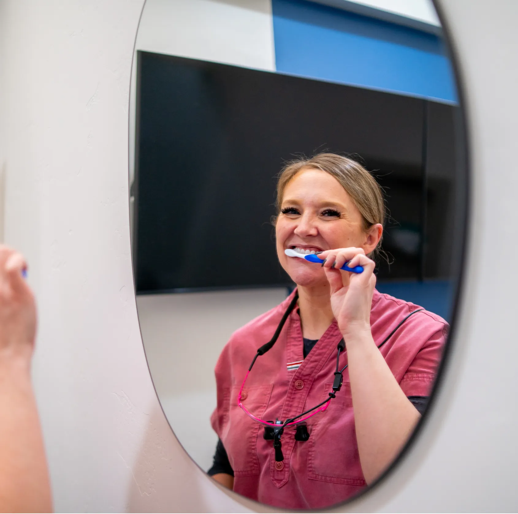Woman in pink dental scrubs brushing her teeth while looking in a mirror.