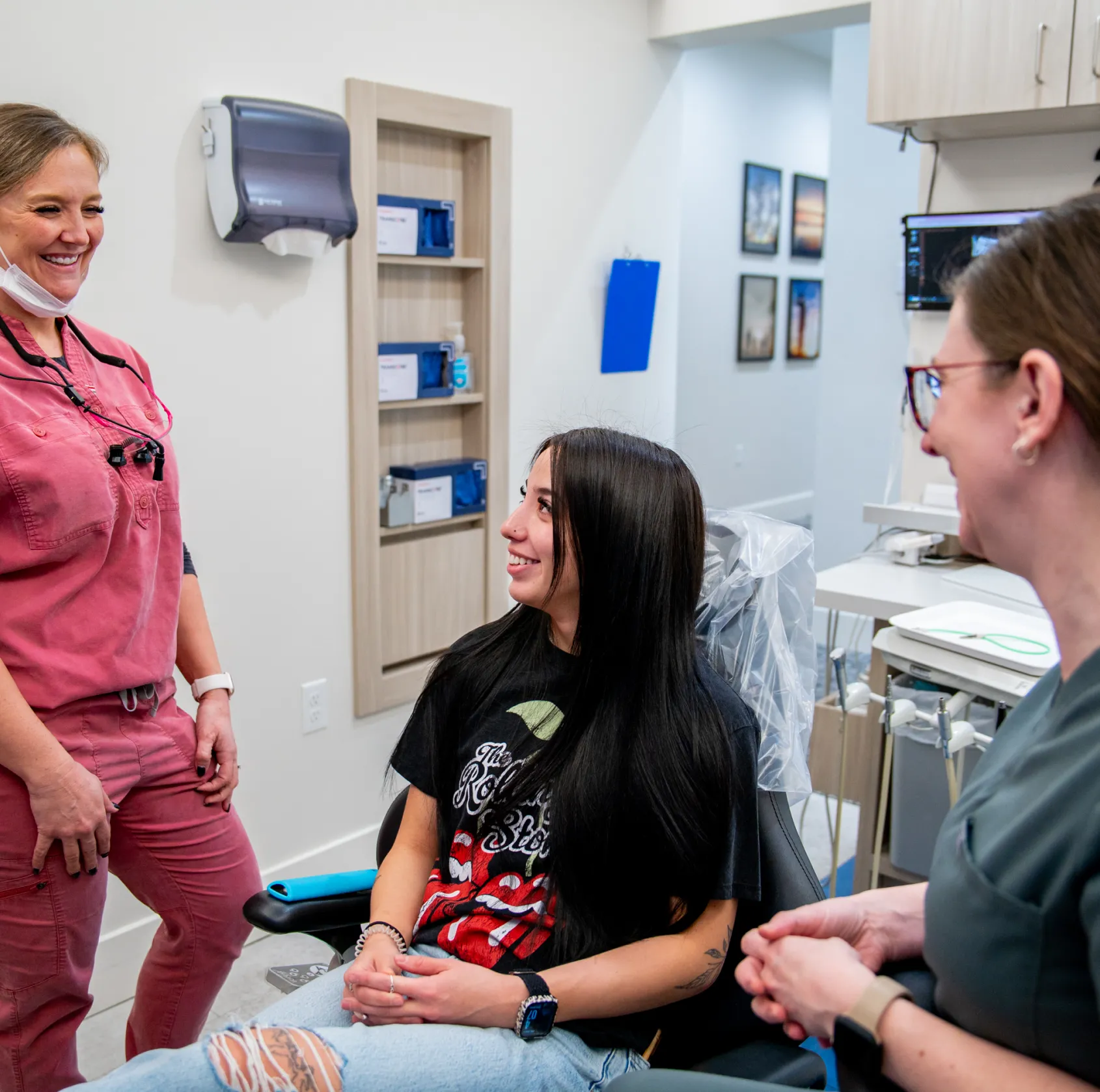 Young woman sitting in a dental chair smiling and talking with two female dental professionals in a dental office.