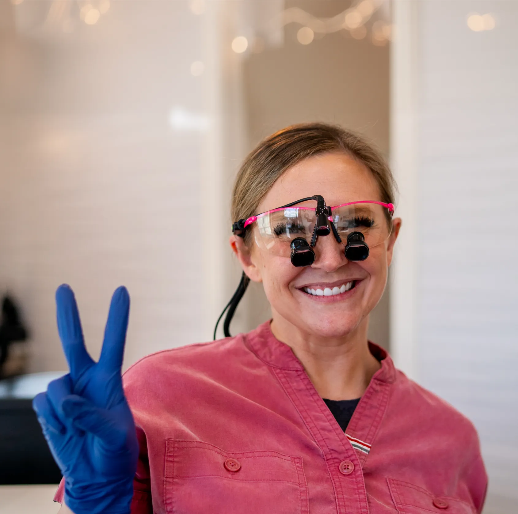 Smiling female dentist wearing magnifying glasses and blue gloves showing a peace sign.