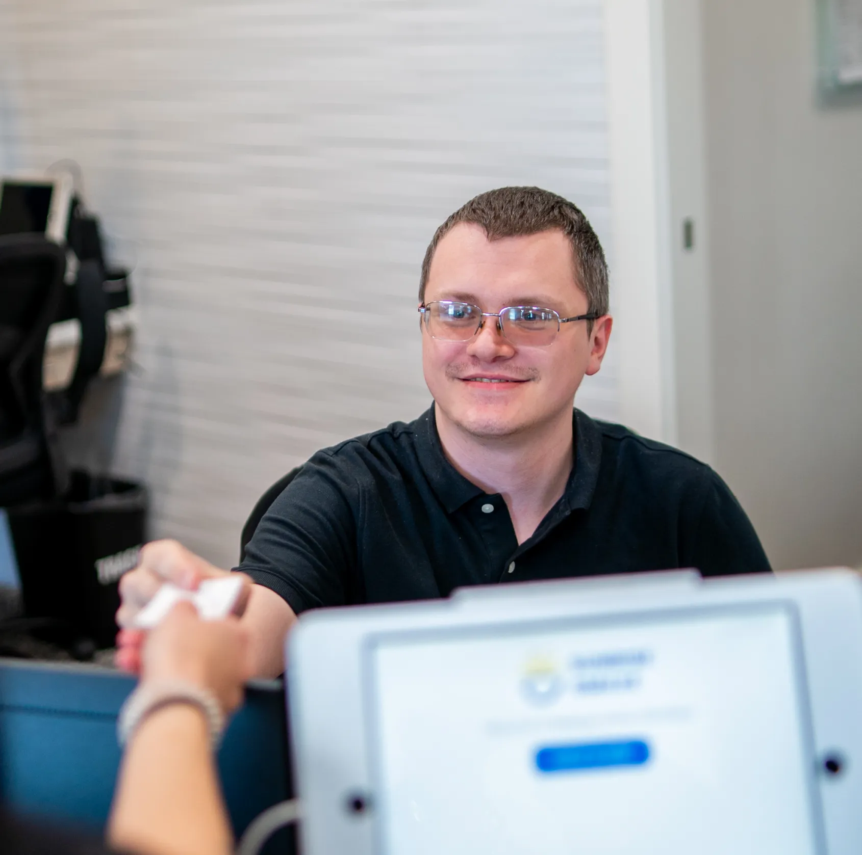 Man wearing glasses and a black polo shirt smiling and handing a card to another person over a desk with a computer screen.