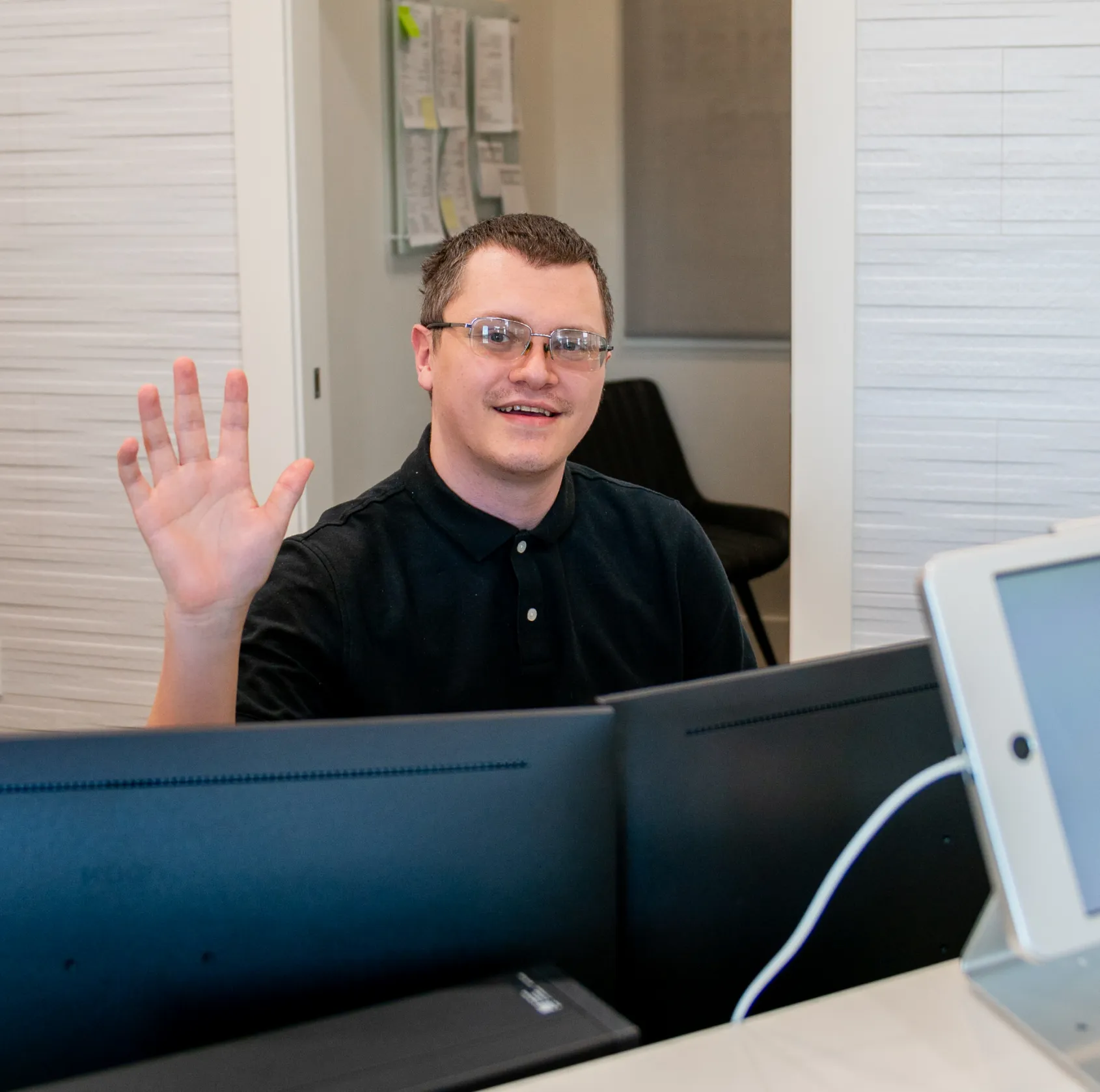 Smiling man wearing glasses waves behind a desk with computer monitors and a tablet in an office setting.