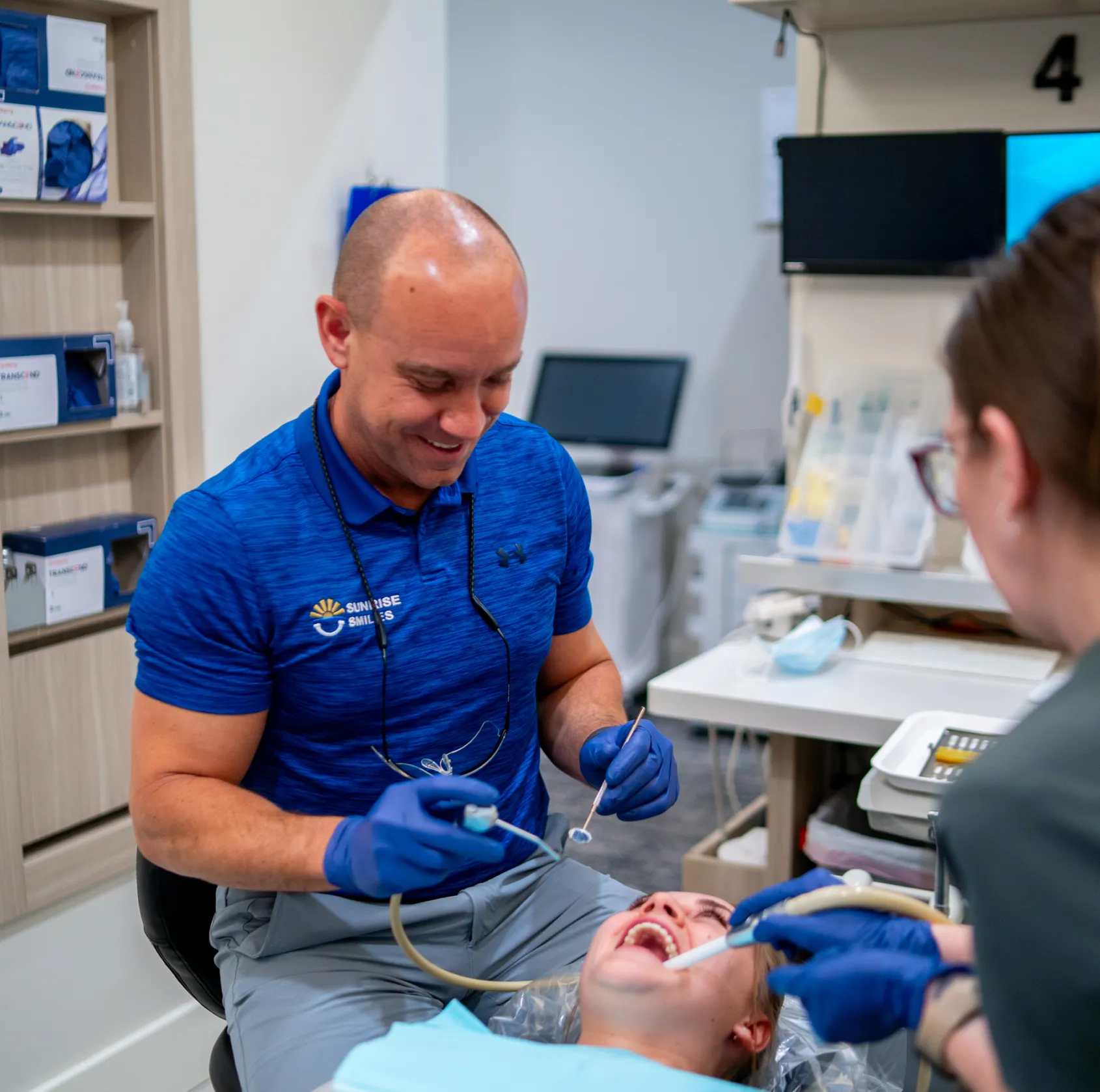 Dentist smiling while performing dental treatment on a patient lying in a dental chair, assisted by a dental hygienist.