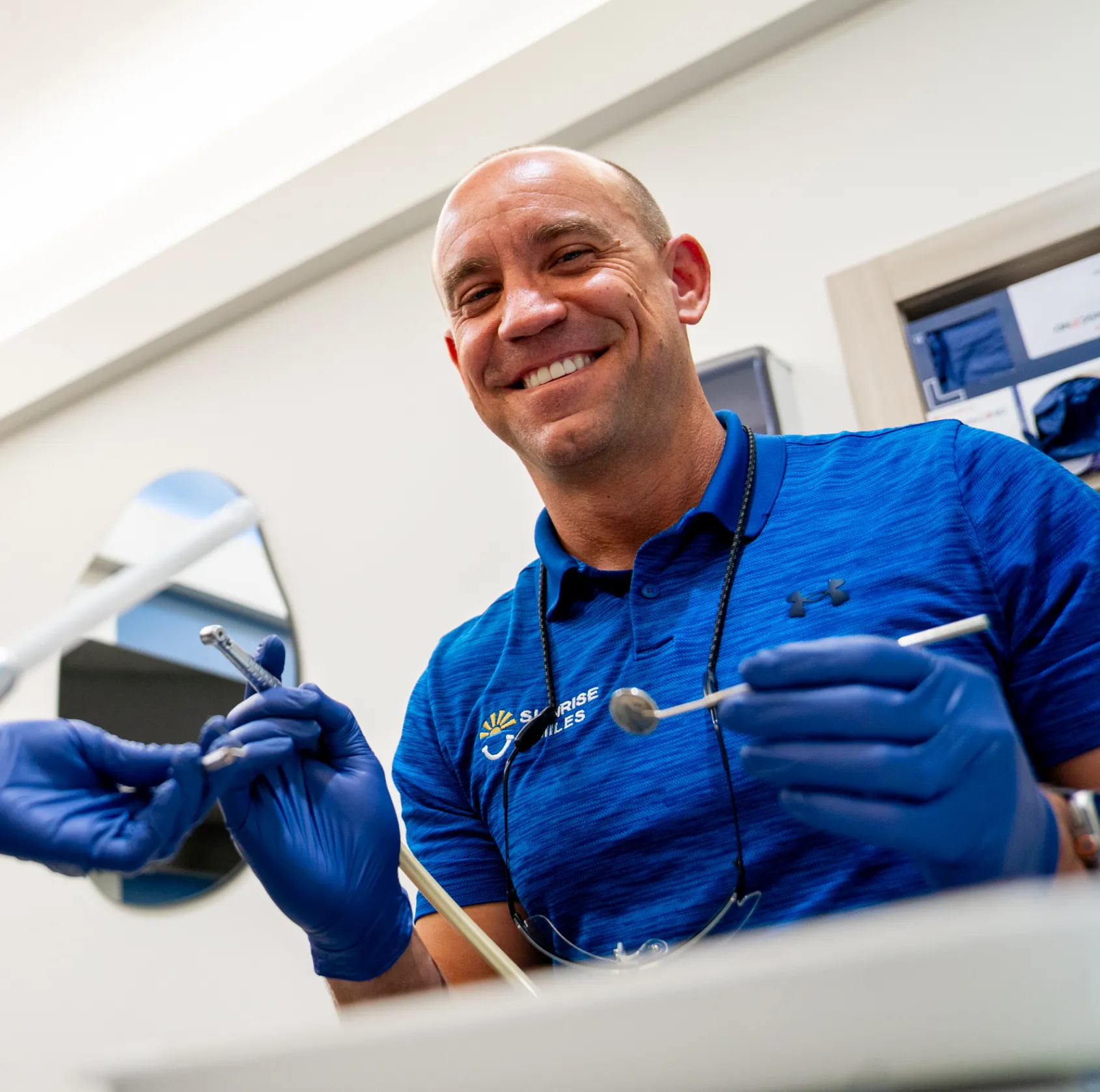 Smiling dentist in blue gloves and a blue shirt holding dental tools in a dental office.