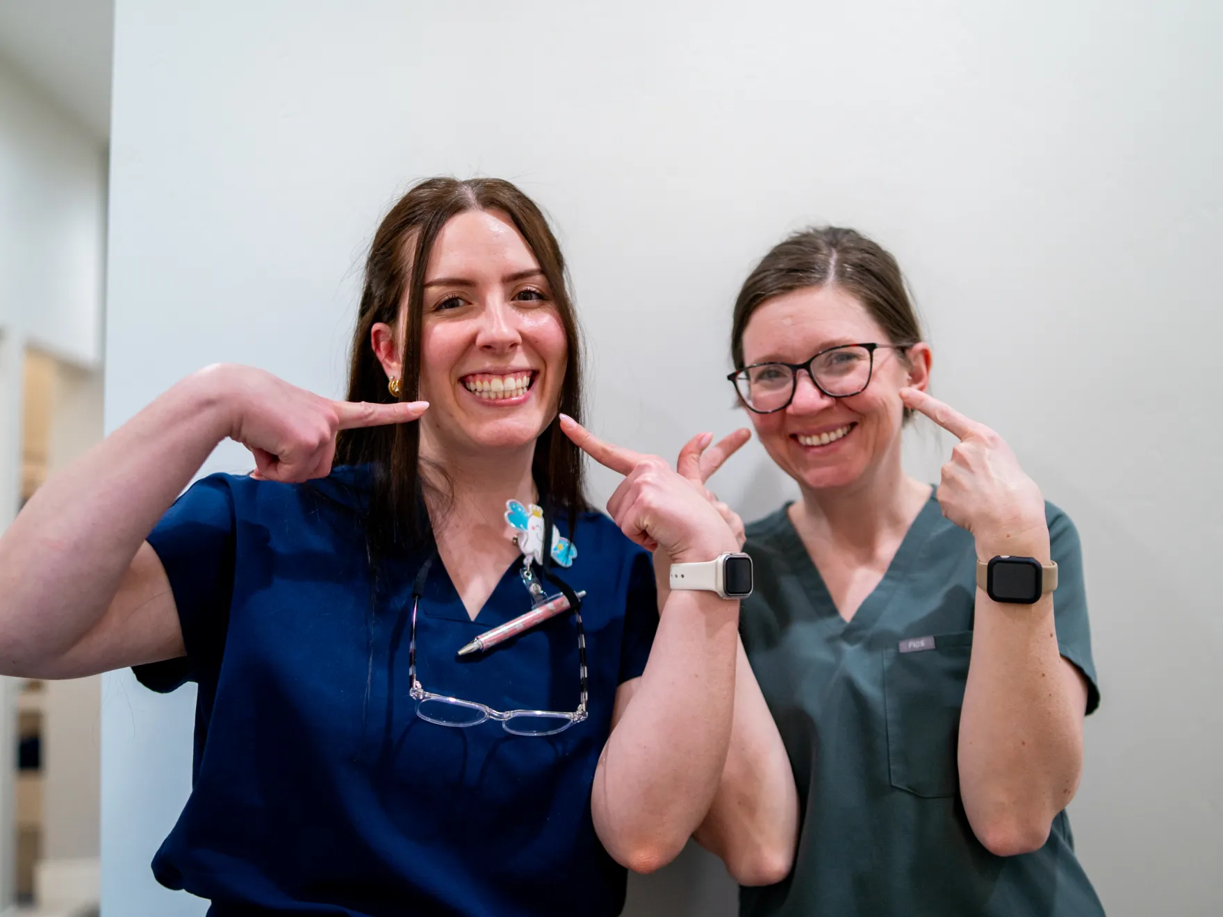 Two smiling female healthcare professionals in scrubs pointing at their teeth.