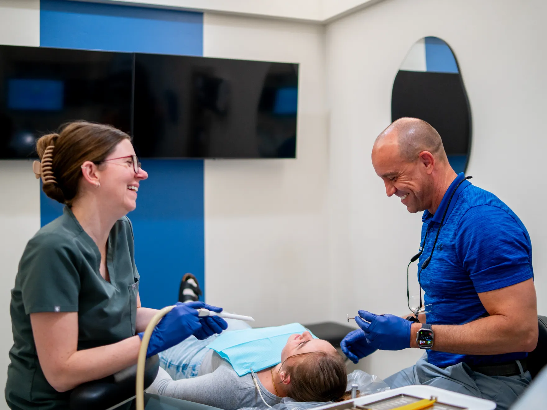 Two dental professionals smiling and attending to a patient lying on the dental chair during a checkup.