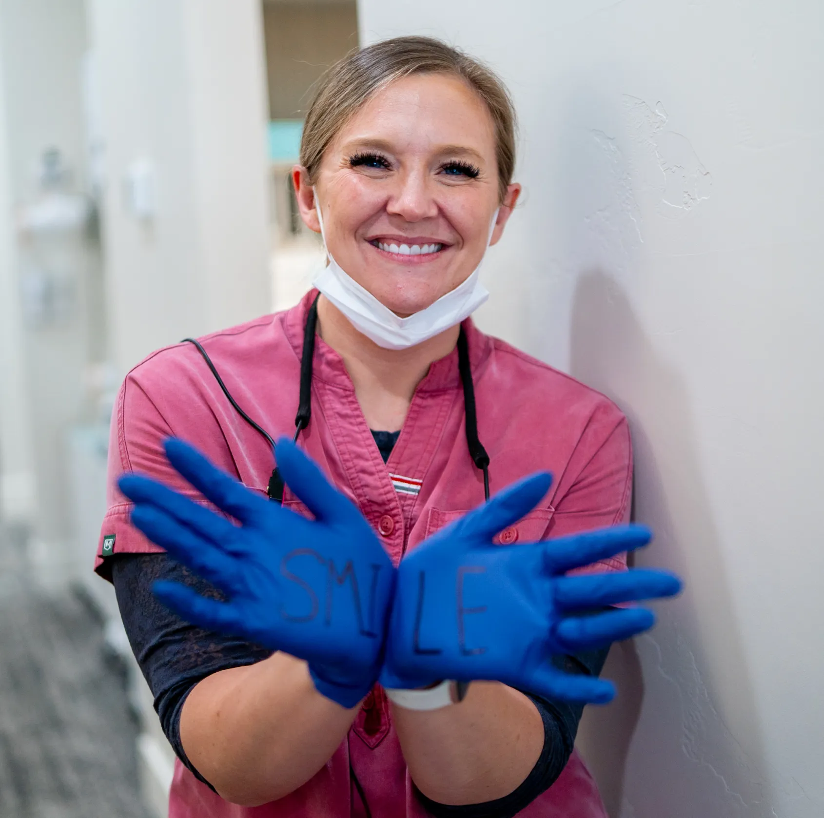 Smiling healthcare worker wearing blue gloves with the word 'SMILE' written on them, a pink scrub top, and a face mask pulled down below the chin.