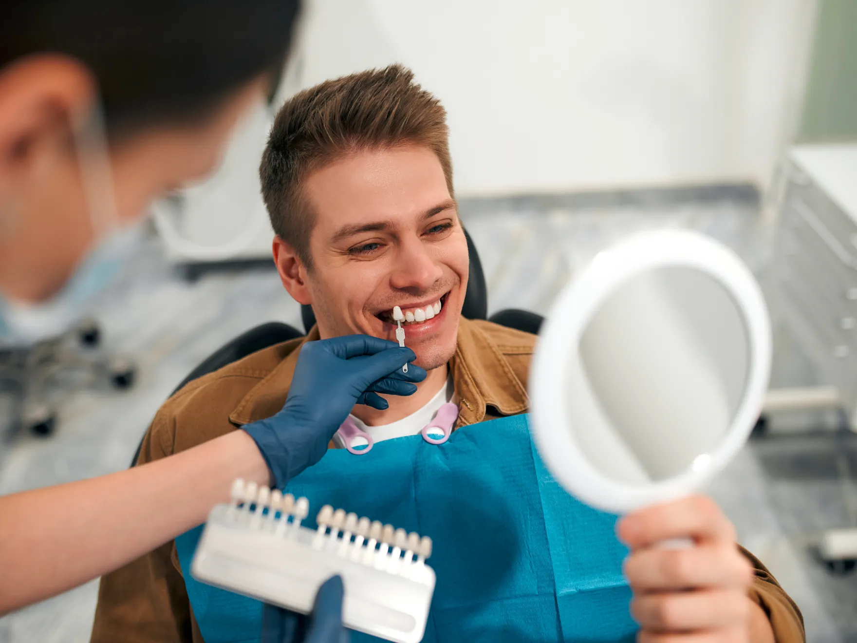 Dentist matching a dental veneer shade to a smiling man's teeth while he holds a mirror.