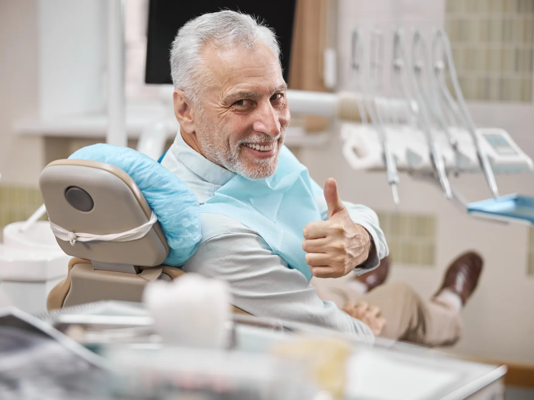 Smiling senior man giving thumbs up while sitting in a dental chair with a protective bib.