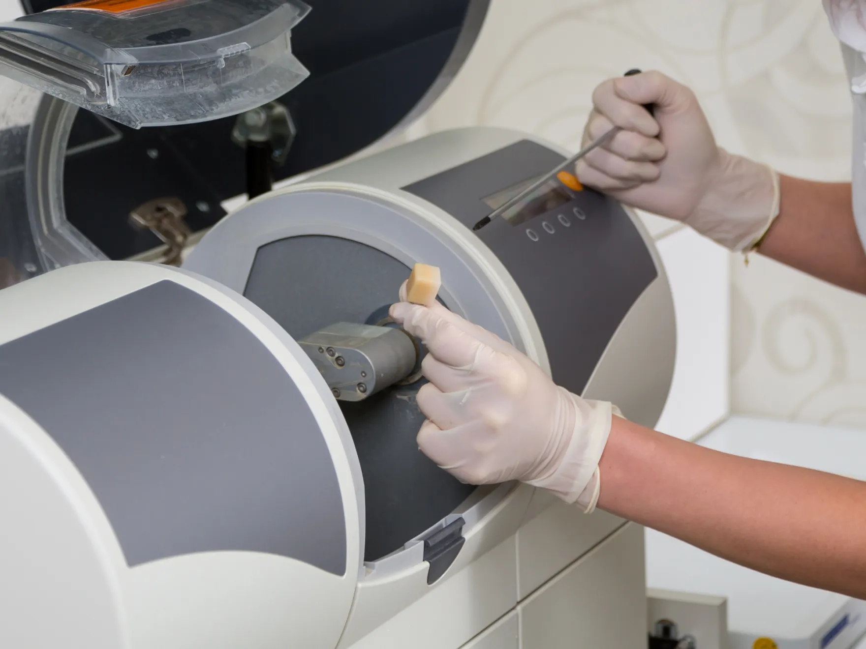 Dental technician wearing gloves using a dental milling machine to shape a dental crown.