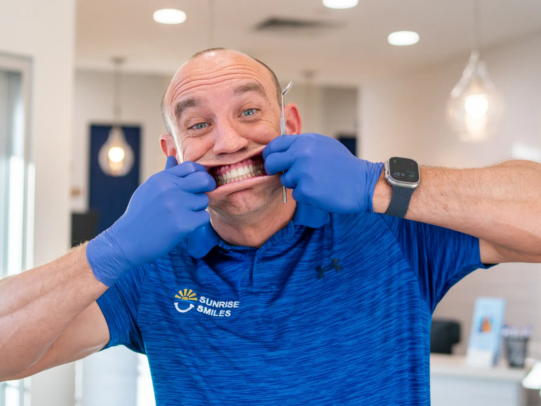 Man wearing blue Sunrise Smiles shirt and gloves pulls back his smile showing clean teeth with a dental tool in his hand.