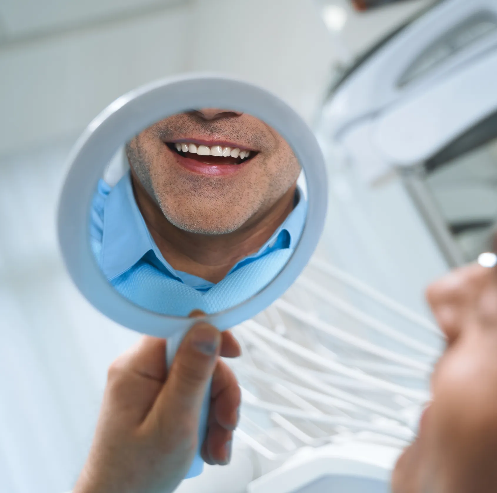 Close-up of a man smiling and looking at his teeth in a handheld mirror at a dental clinic.