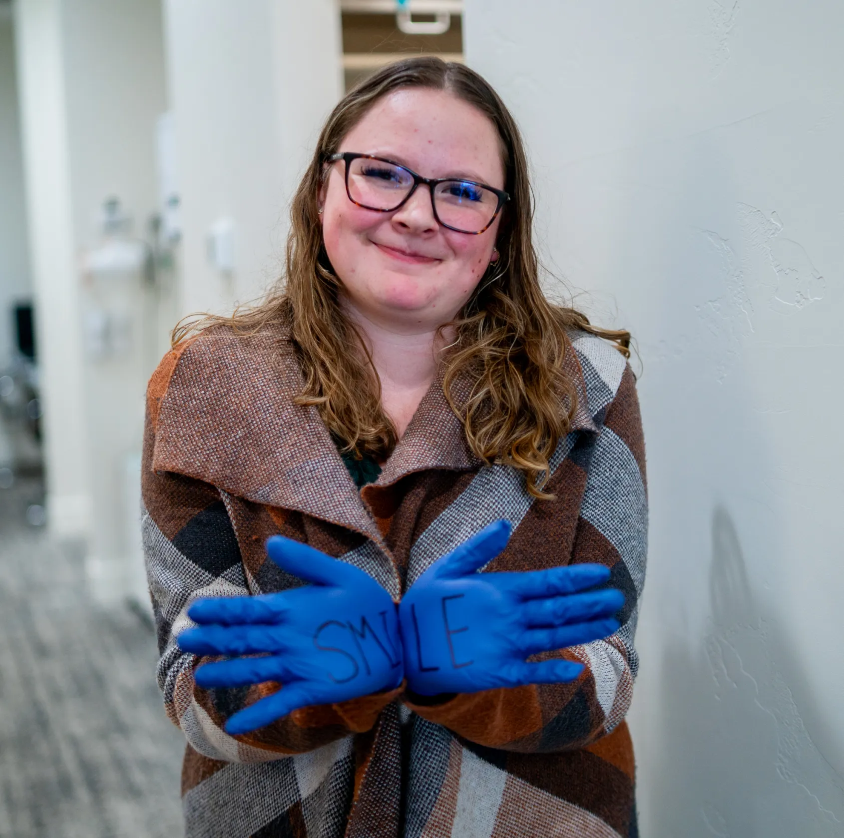 Smiling woman wearing glasses and a plaid coat with blue gloves showing the word 'SMILE' written across her palms.