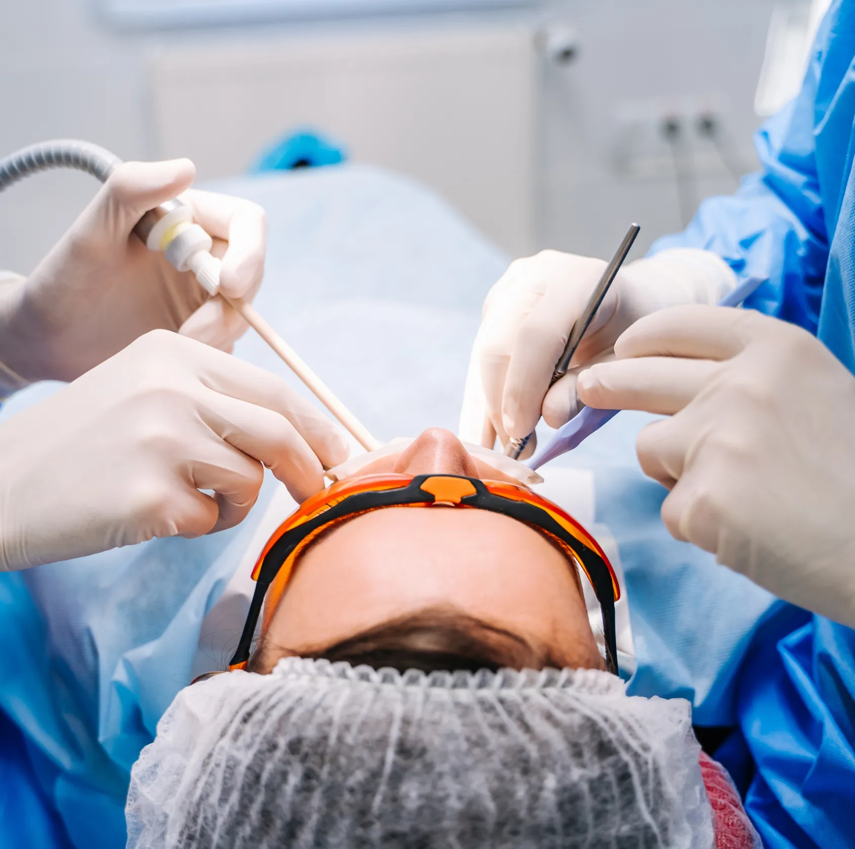 Patient wearing orange protective glasses undergoing dental treatment by gloved professionals using dental instruments.