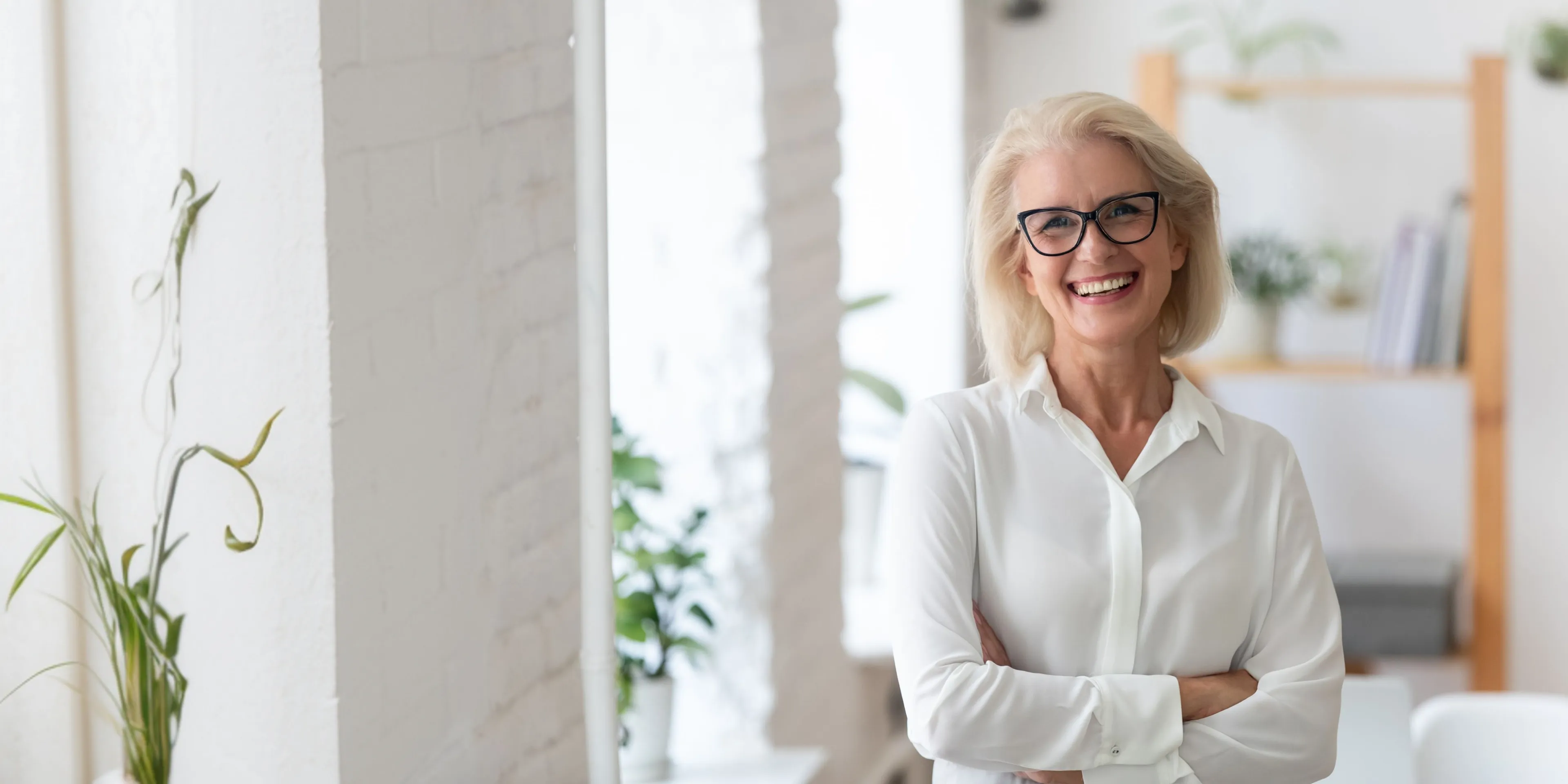 Smiling mature woman with glasses wearing a white shirt standing with arms crossed in a bright modern office.