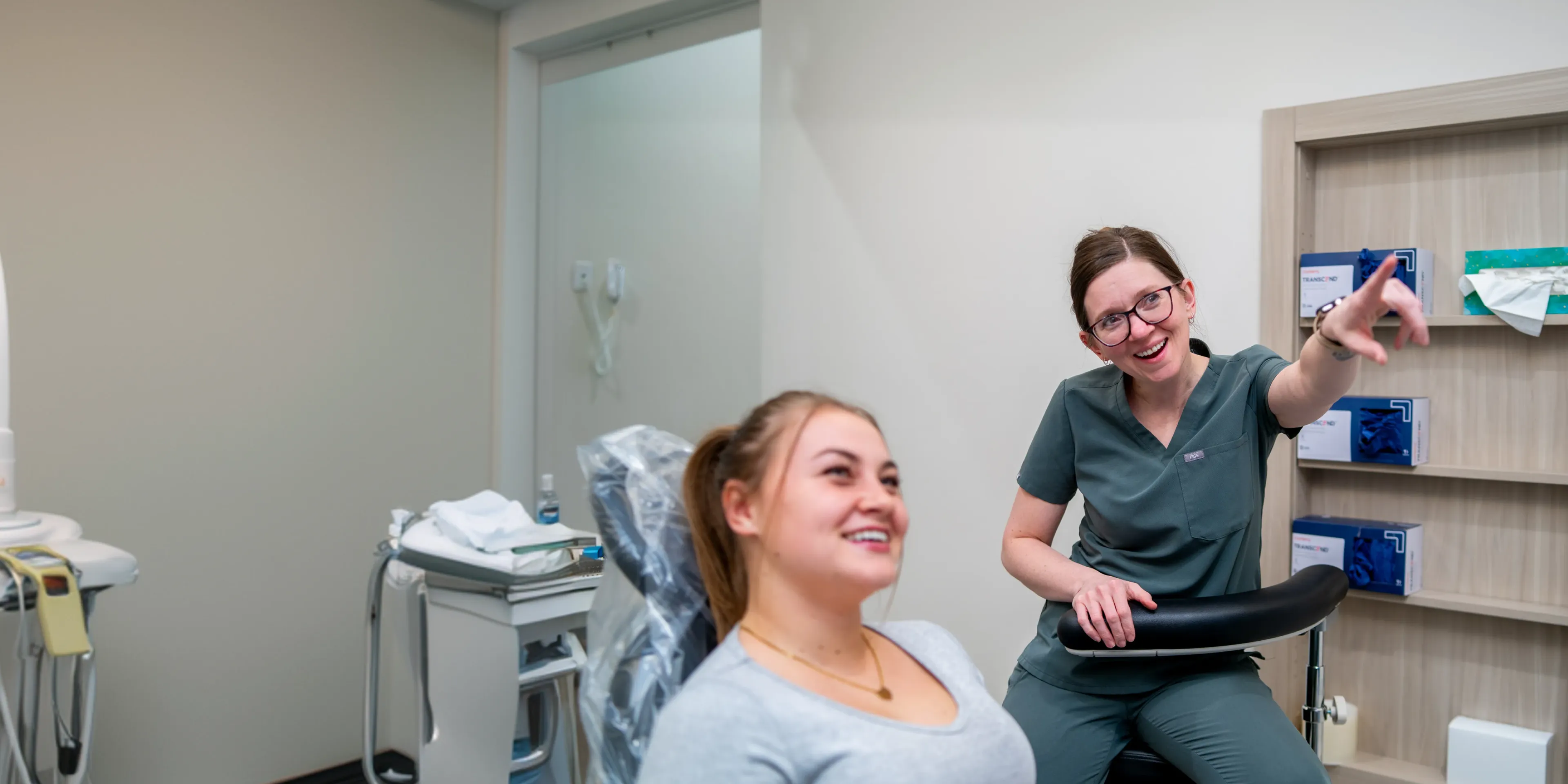 Female medical professional in scrubs pointing and smiling while talking to a female patient in a clinic room.