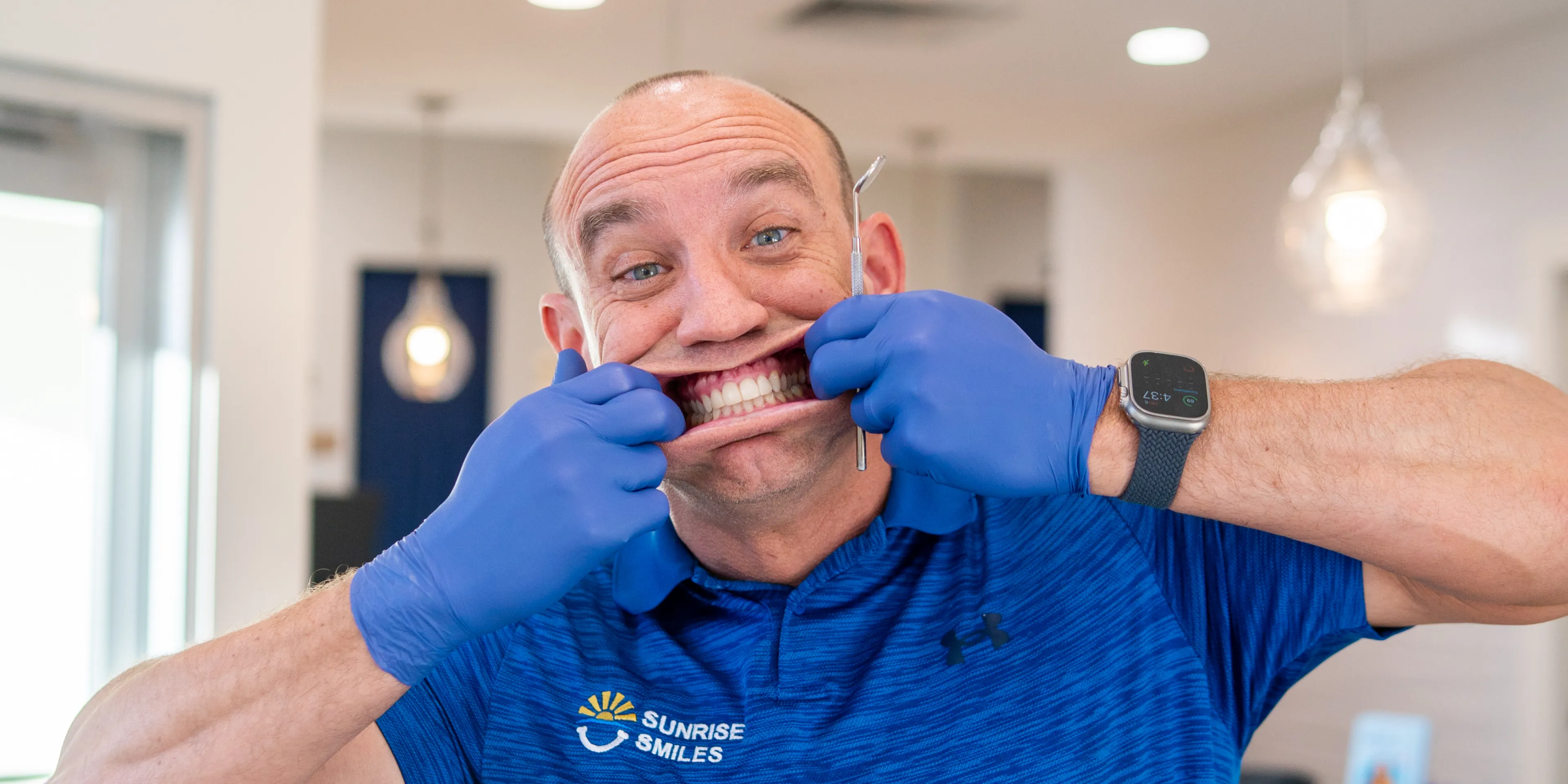 Smiling man wearing blue gloves and a Sunrise Smiles shirt stretches his mouth to show teeth holding a dental tool.