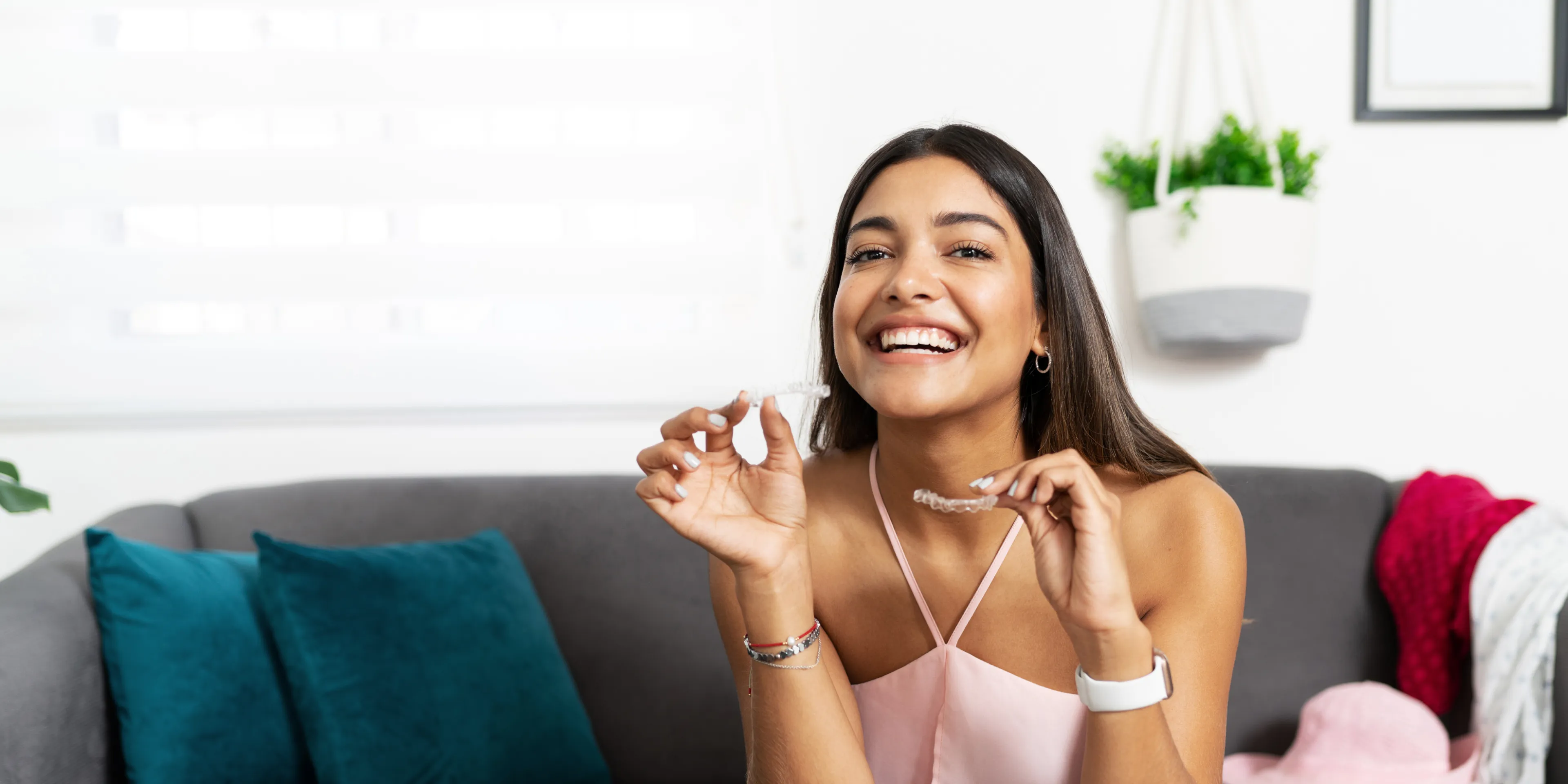 Smiling woman sitting on a gray couch holding clear Invisalign aligners in both hands.