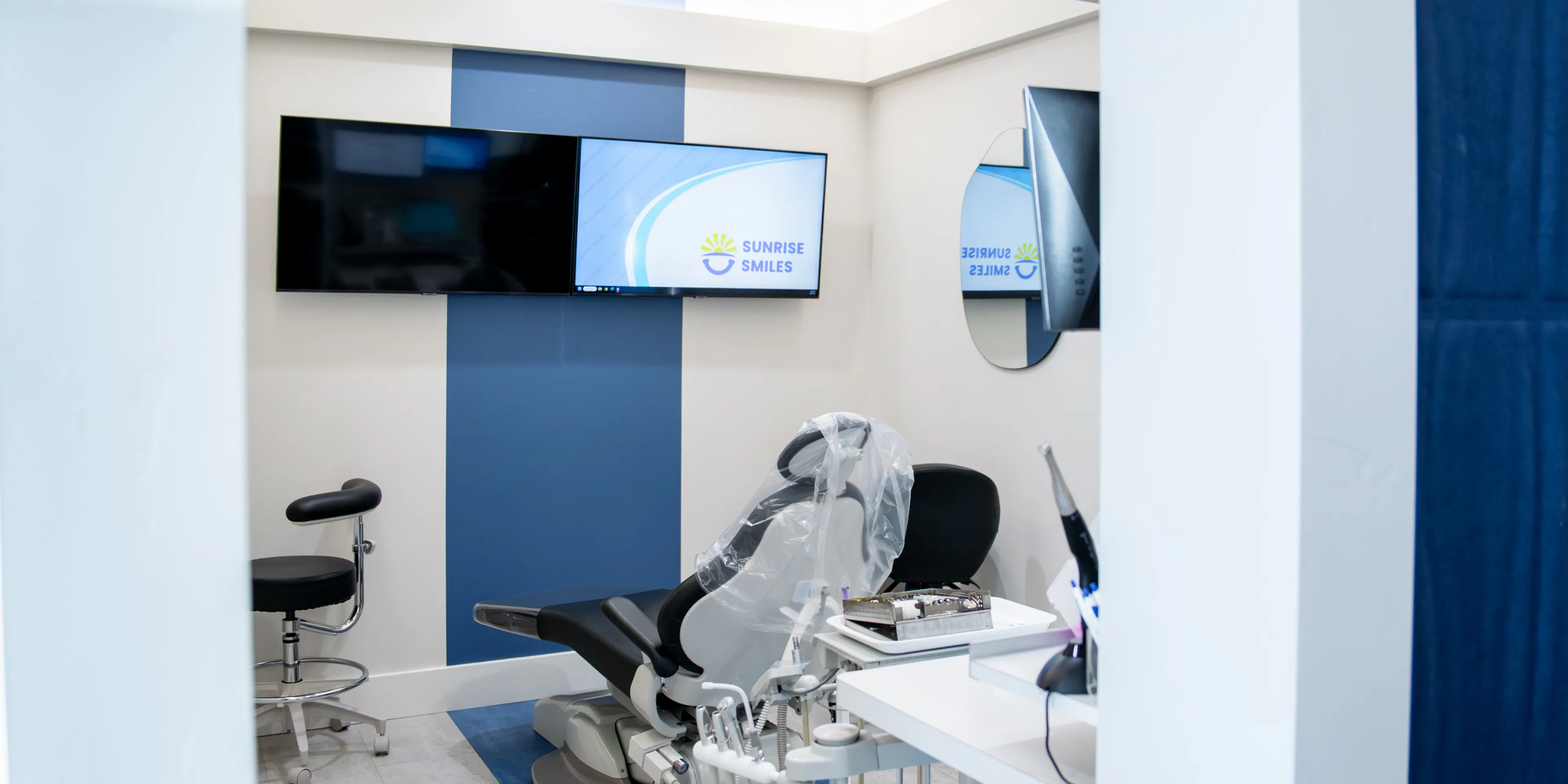 Modern dental clinic room with a dental chair covered in plastic, medical tools, and two mounted flat-screen monitors one showing Sunrise Smiles logo.