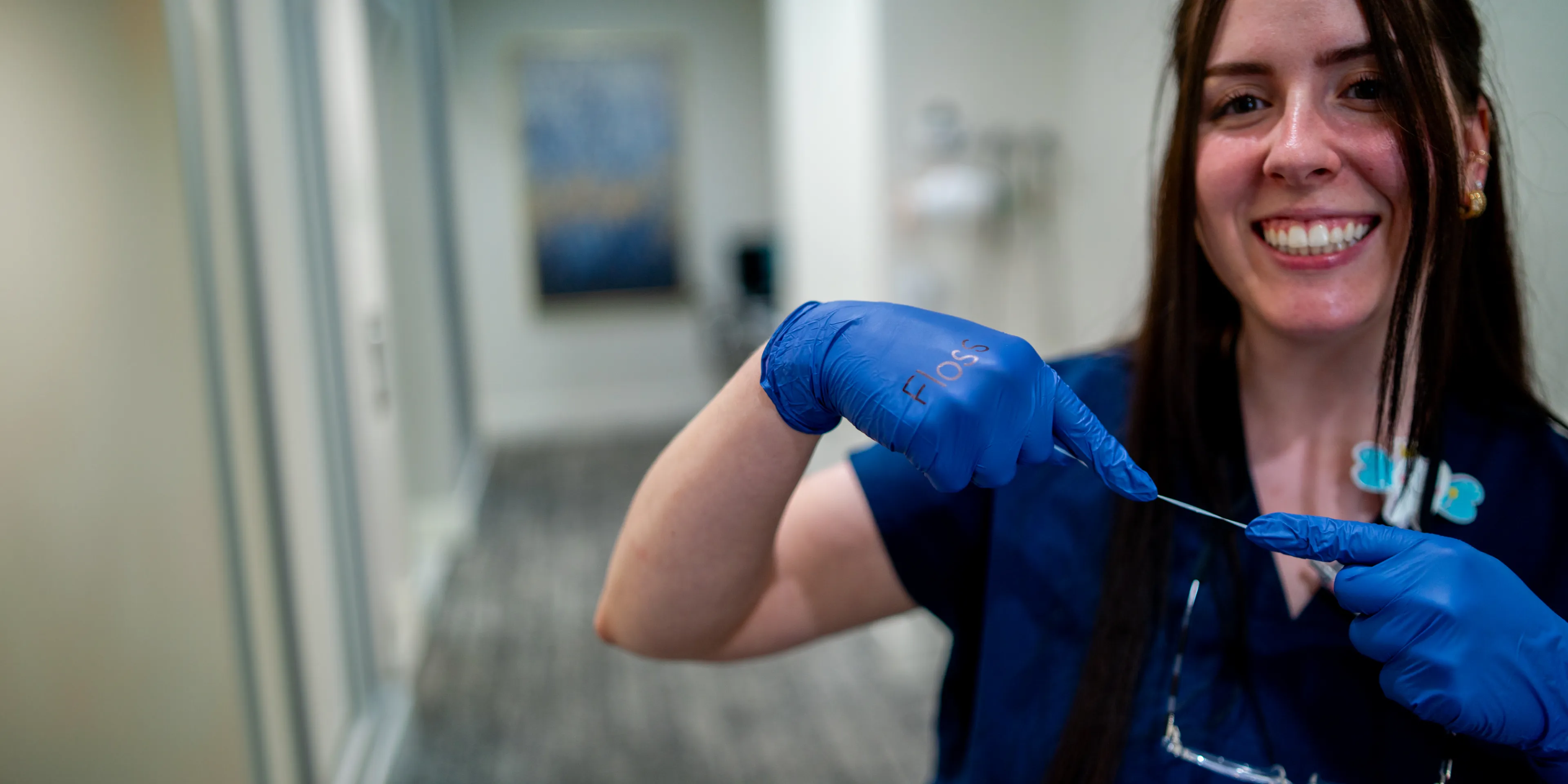 Smiling dental professional wearing blue gloves holding a piece of dental floss stretched between fingers.