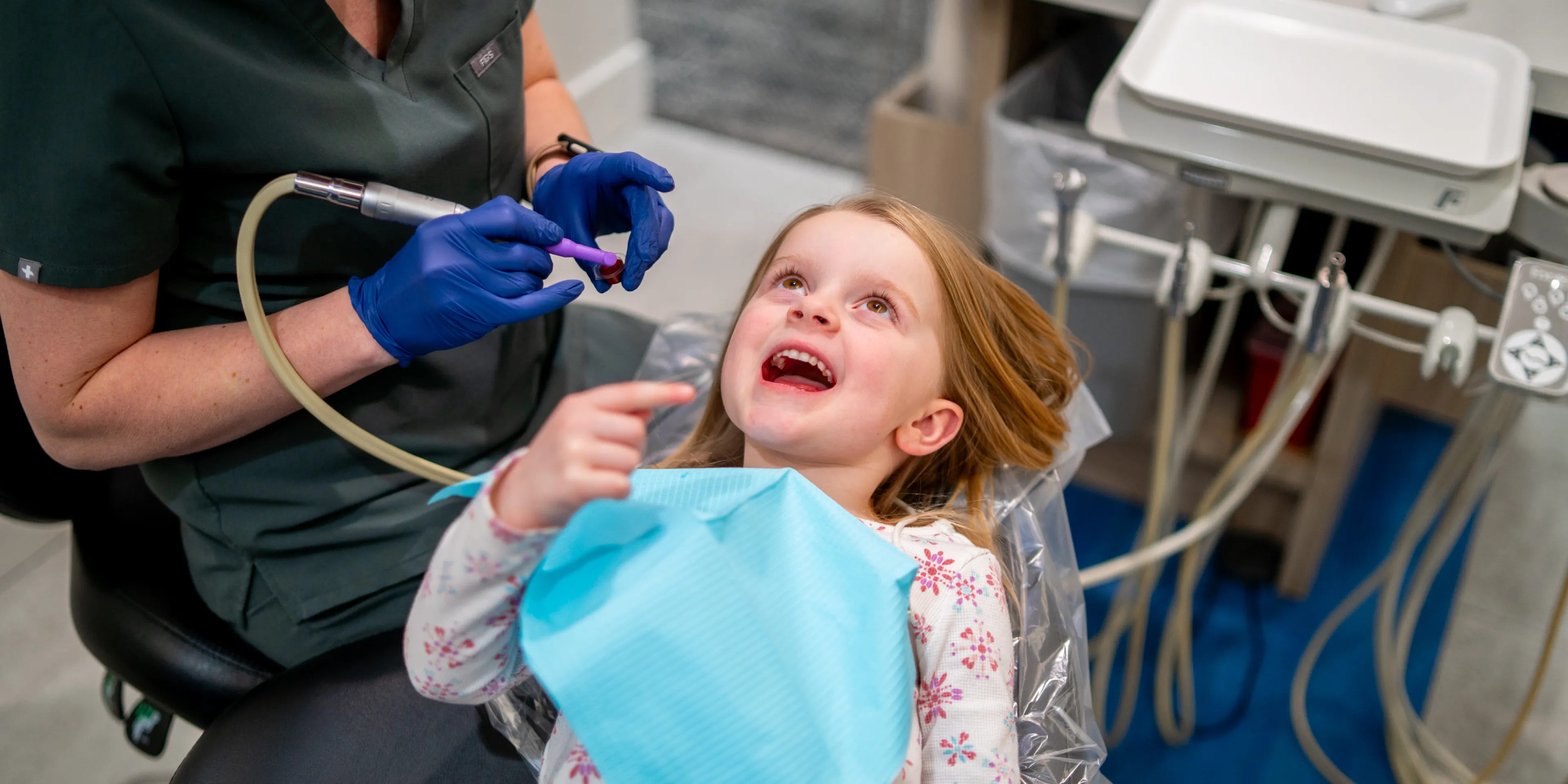 Young girl sitting in a dental chair with a dental bib pointing inside her mouth as a dental professional wearing gloves prepares a dental tool.