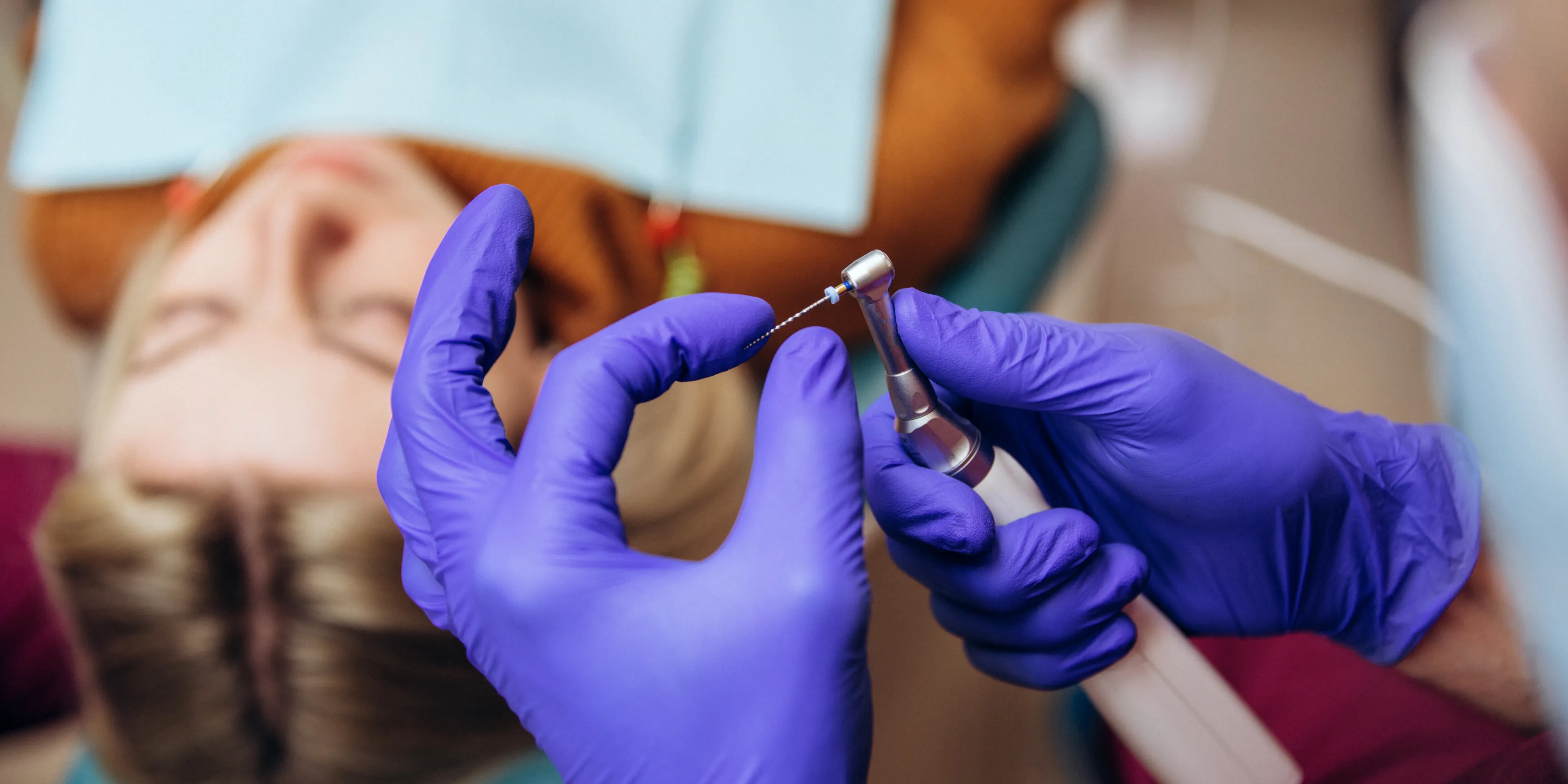 Close-up of a dentist's gloved hands holding a dental tool with a small rotating file, with a patient lying back in the dental chair in the background.