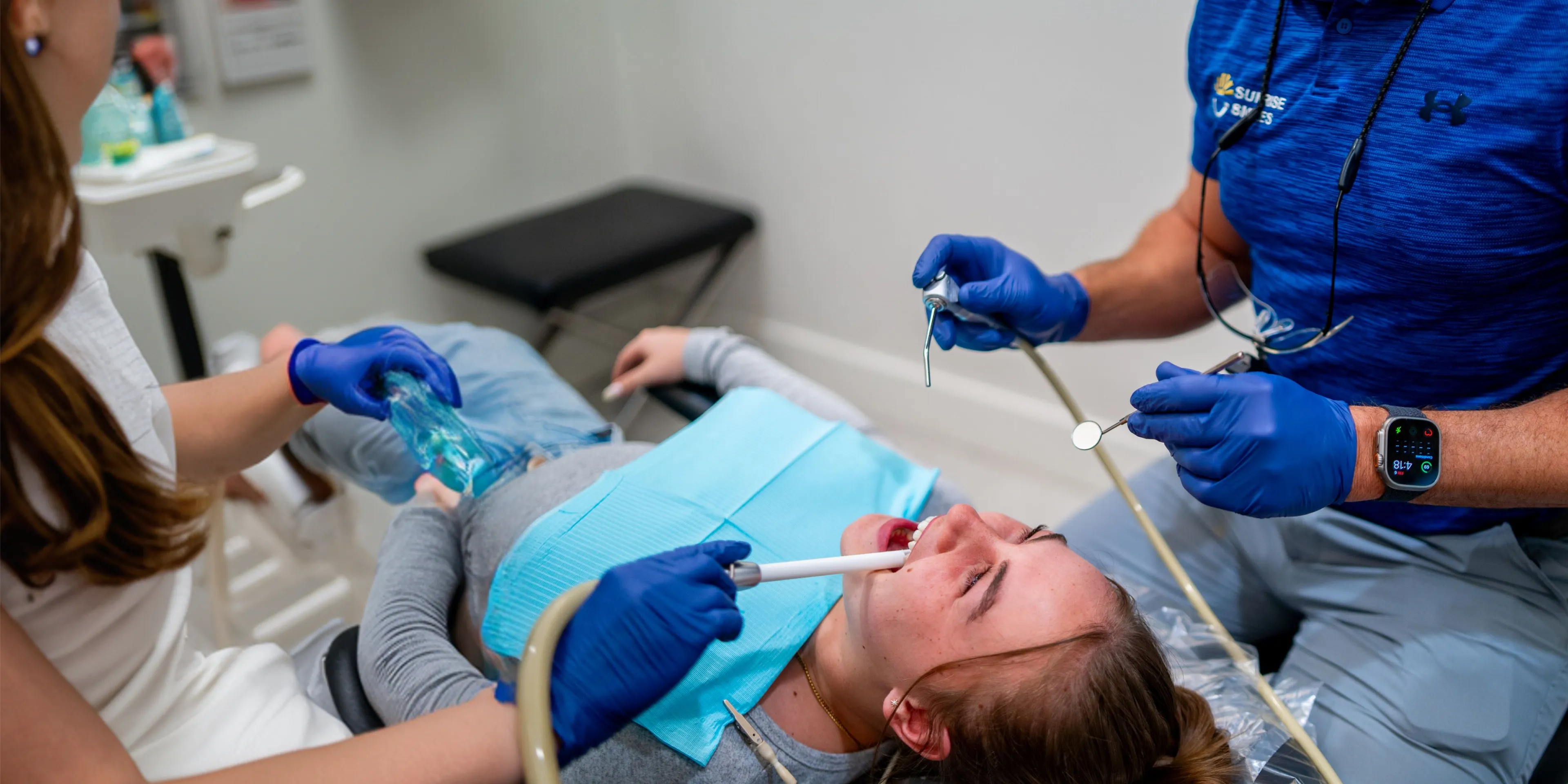 Patient lying in a dental chair receiving treatment from two dental professionals wearing blue gloves.