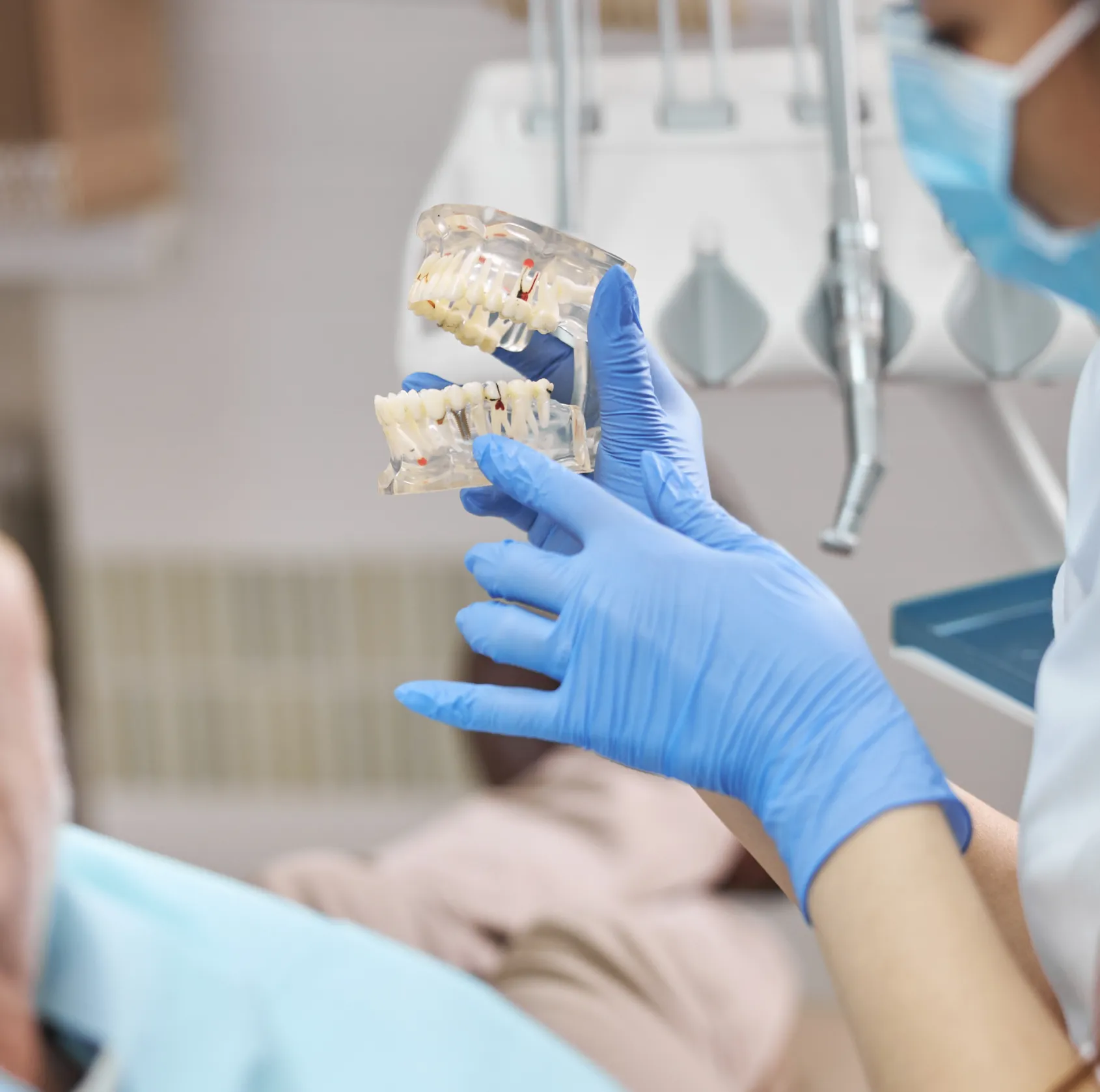 Dentist wearing blue gloves holding a dental model with implants, demonstrating to a patient in a dental office.