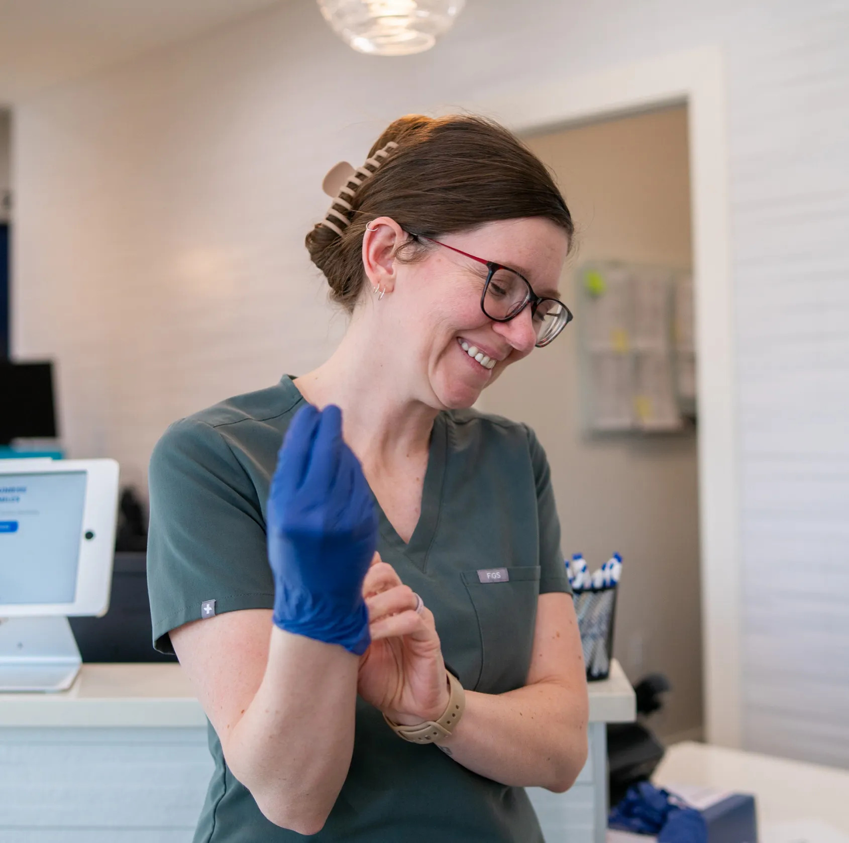 Smiling female healthcare worker wearing scrubs and glasses putting on a blue glove in a medical office.