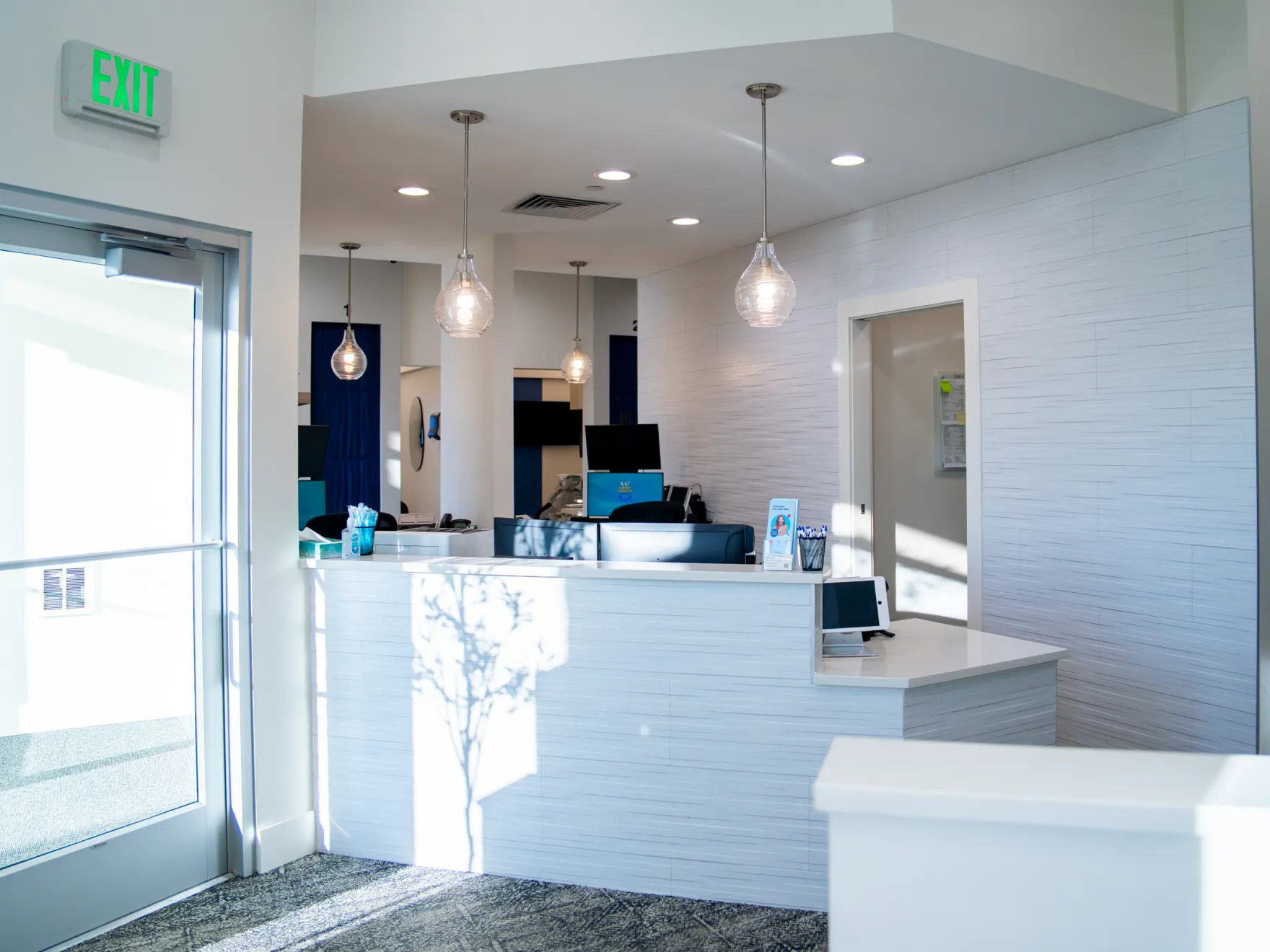 Bright reception area with white desk, hanging pendant lights, and glass door with exit sign.