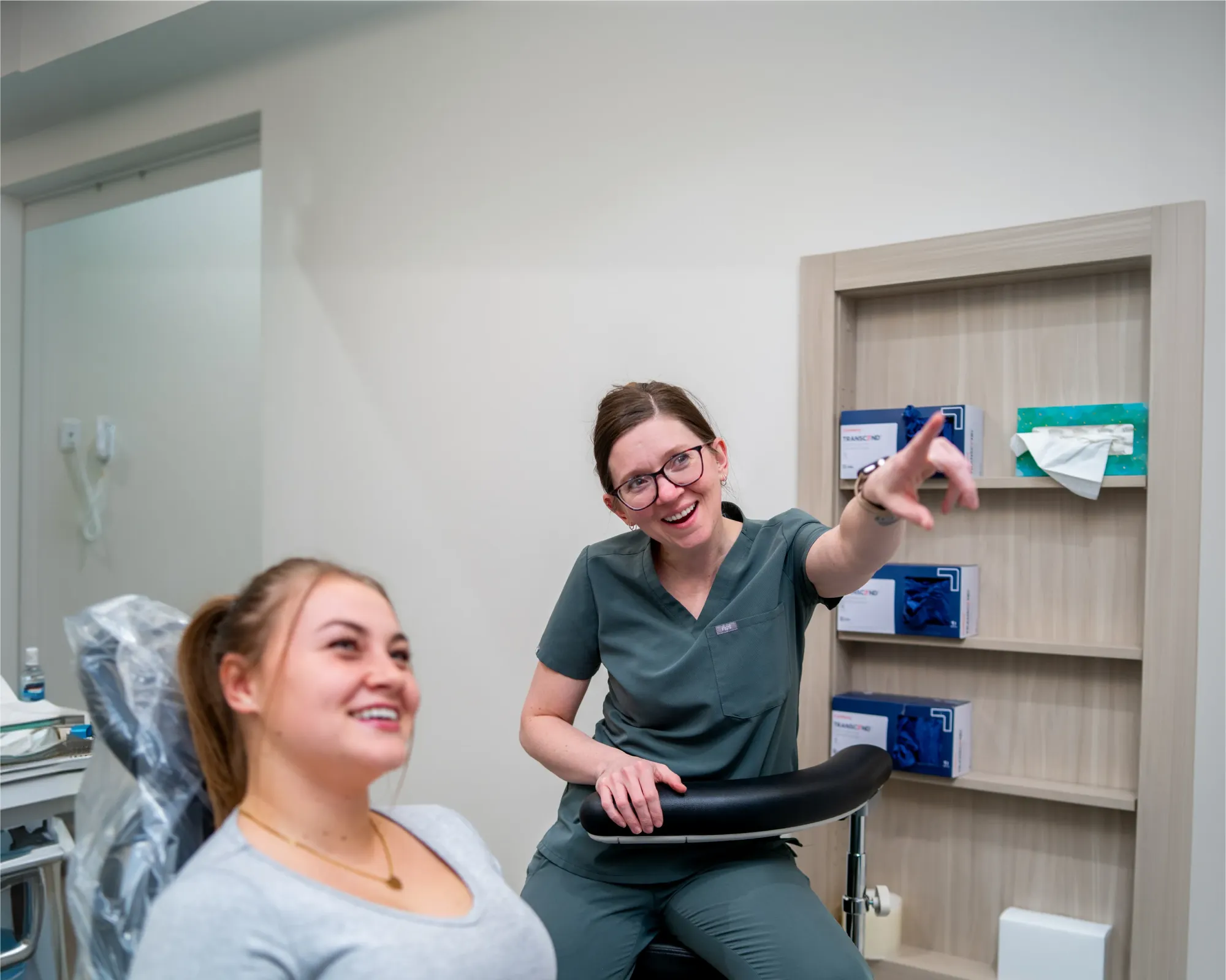 A smiling female healthcare professional in scrubs gestures while talking to a female patient seated in a medical chair.