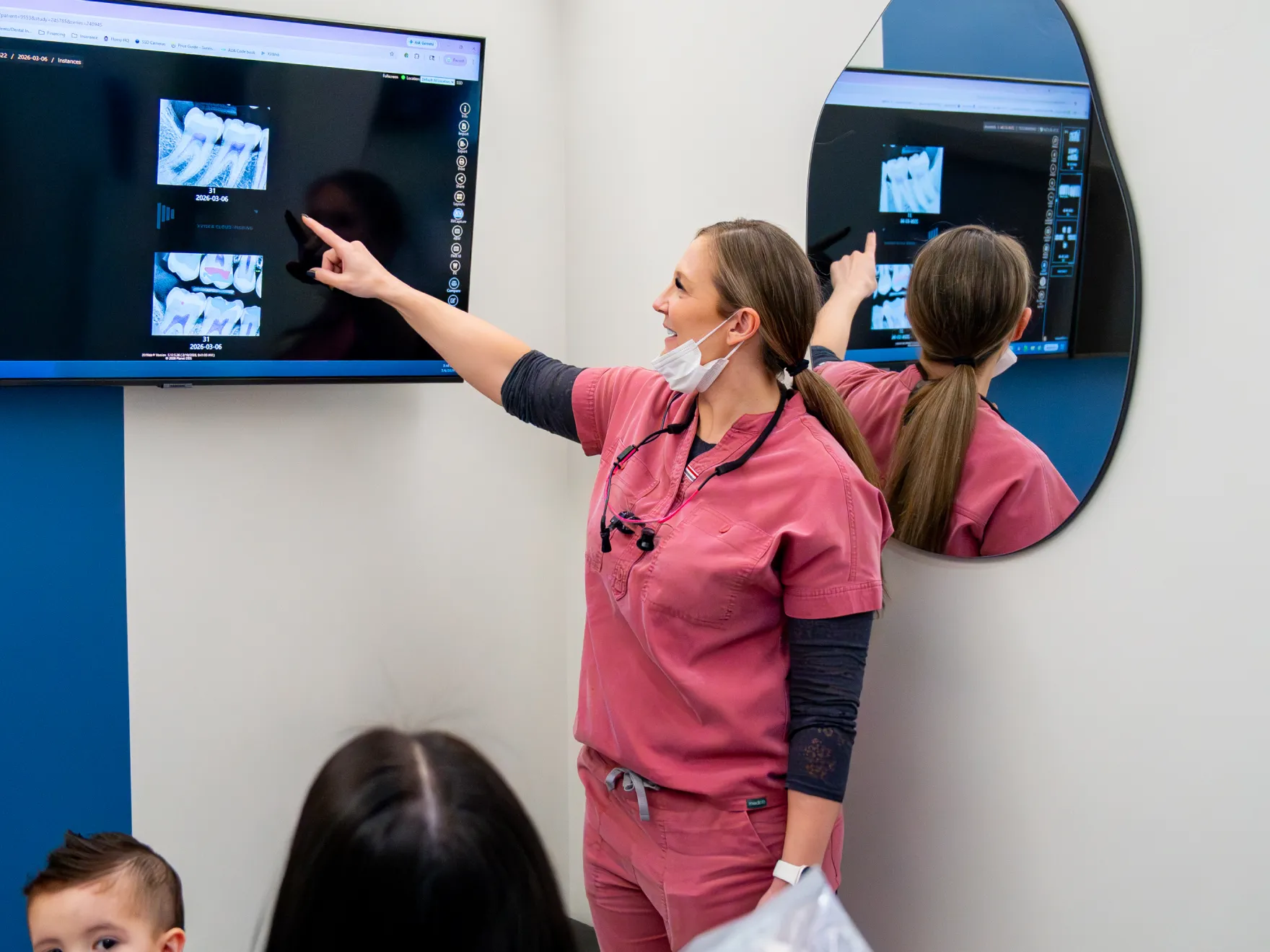 Dental professional in pink scrubs pointing to dental X-rays displayed on a screen in a clinic room.