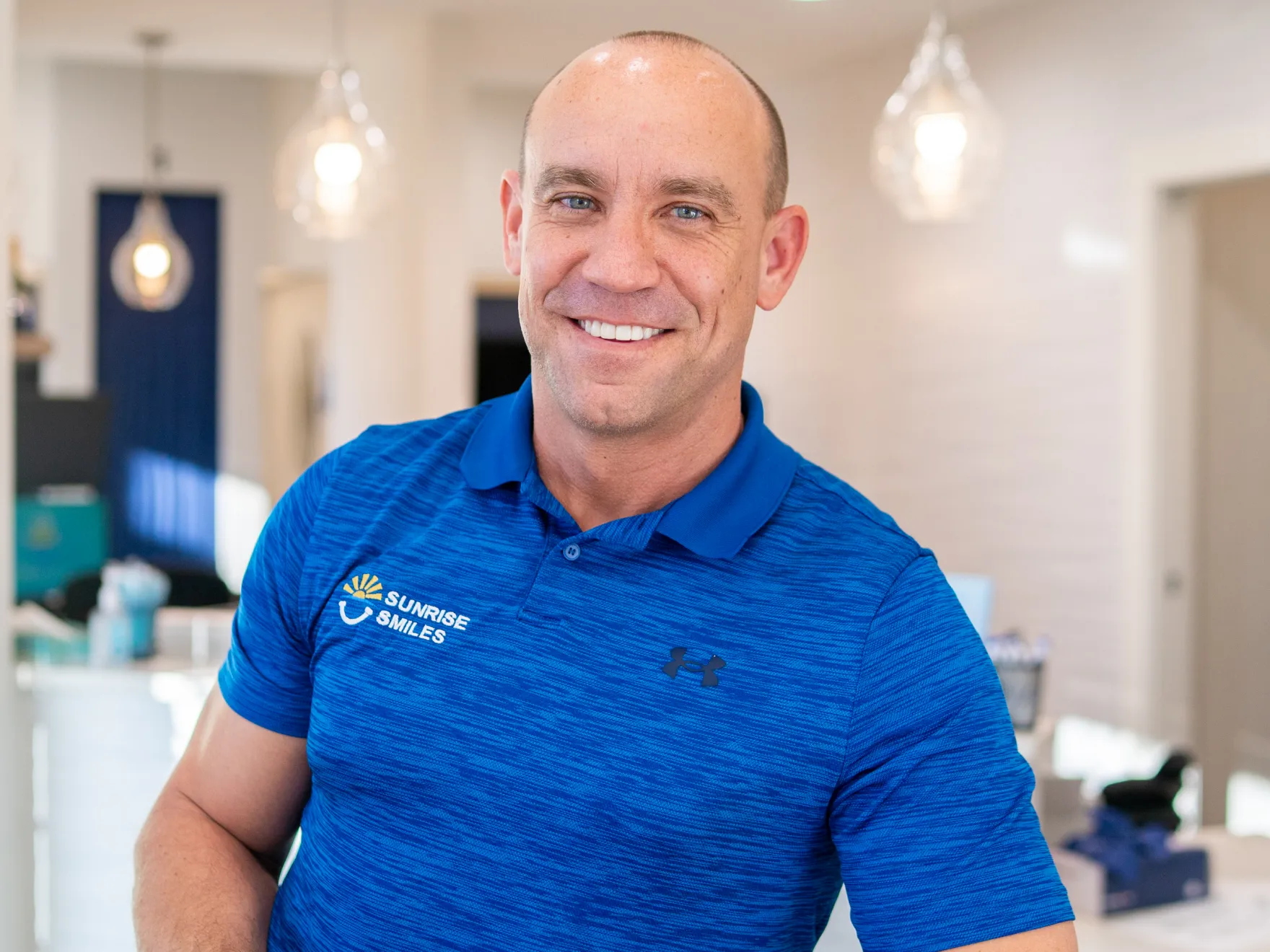 Smiling bald man wearing a blue Sunrise Smiles polo shirt in a bright indoor setting.