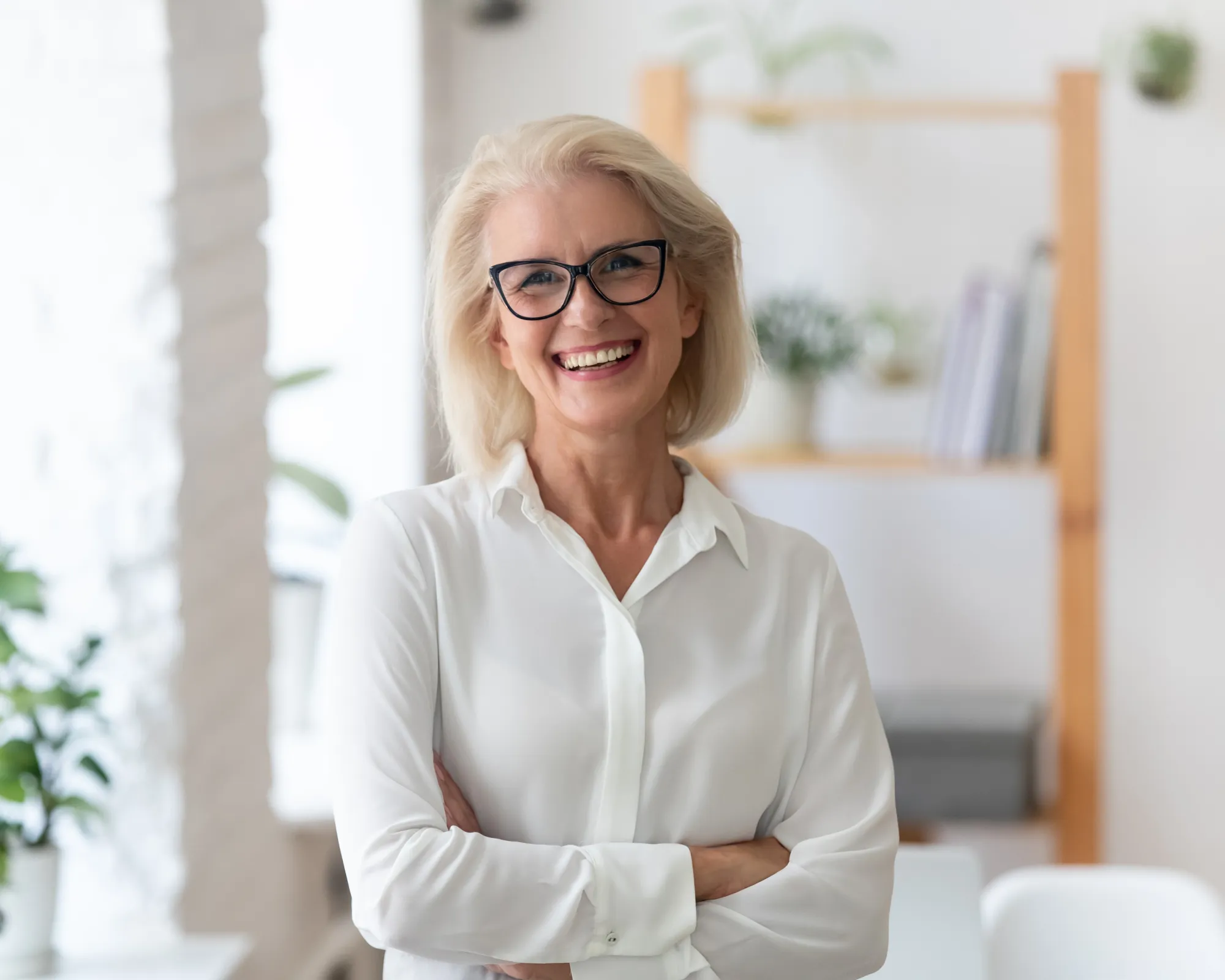 Smiling elderly woman with white hair and glasses wearing a white blouse, standing with arms crossed in a bright, modern office.
