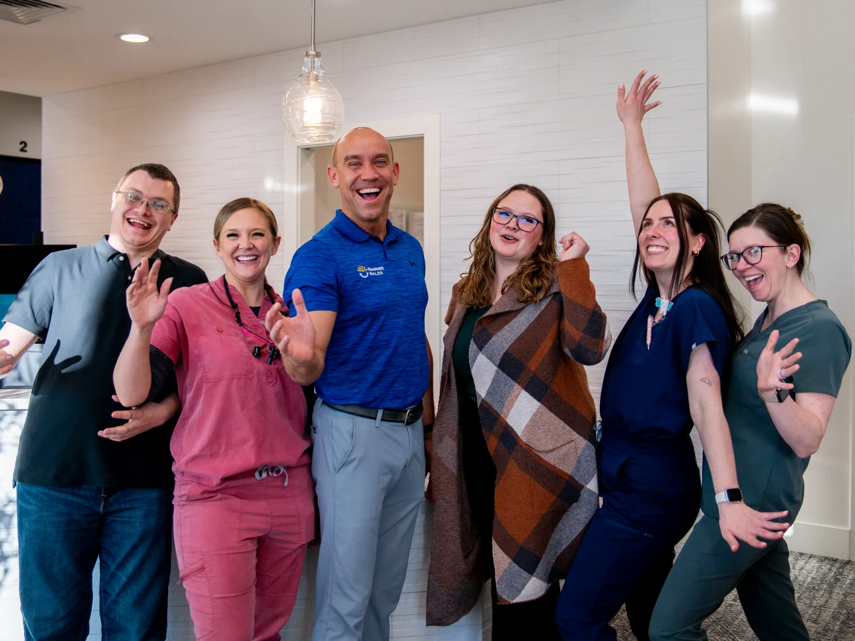 Six cheerful adults standing indoors, smiling and gesturing energetically at the camera.