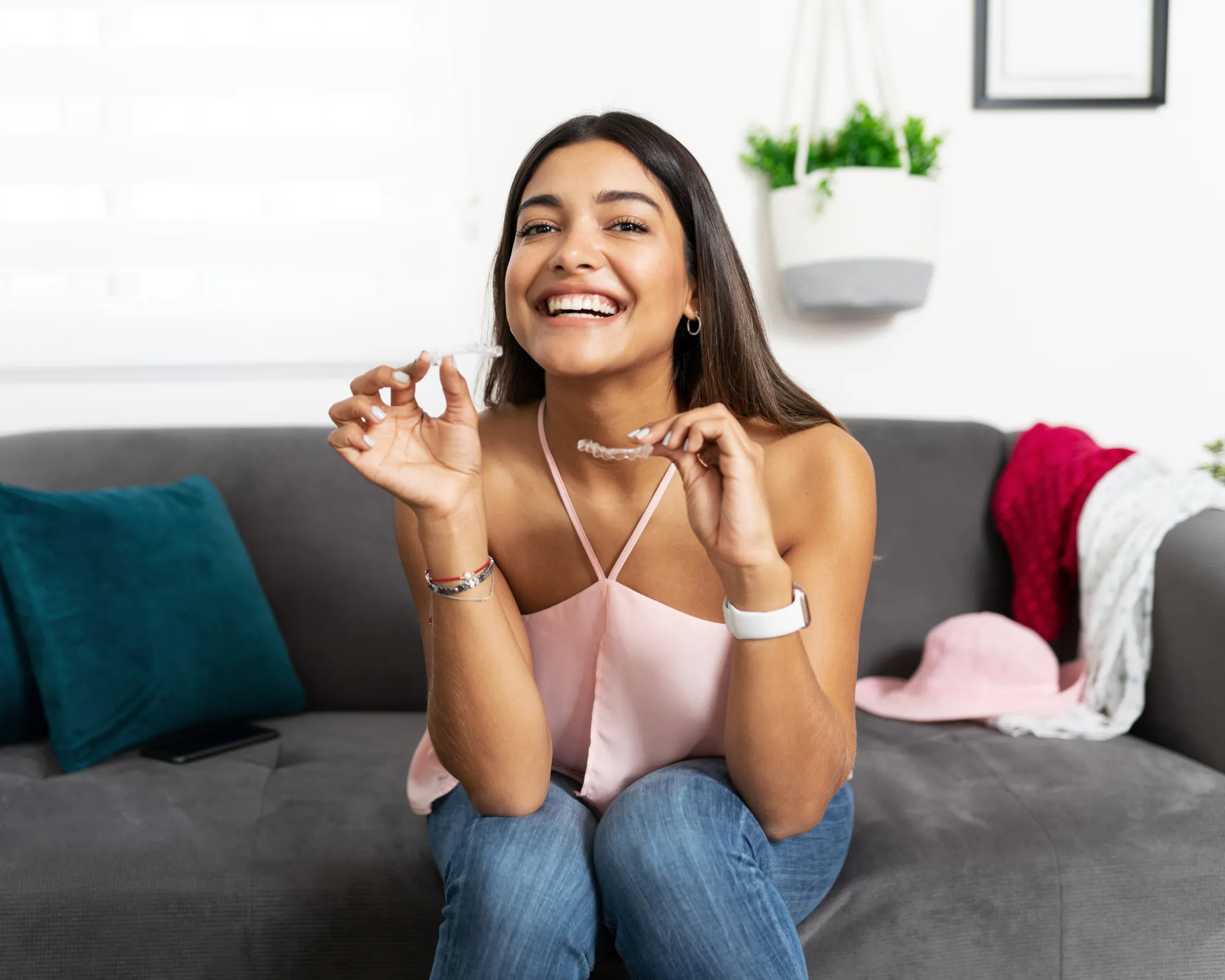 Smiling woman sitting on a couch holding two clear dental aligners, showing them to the camera.