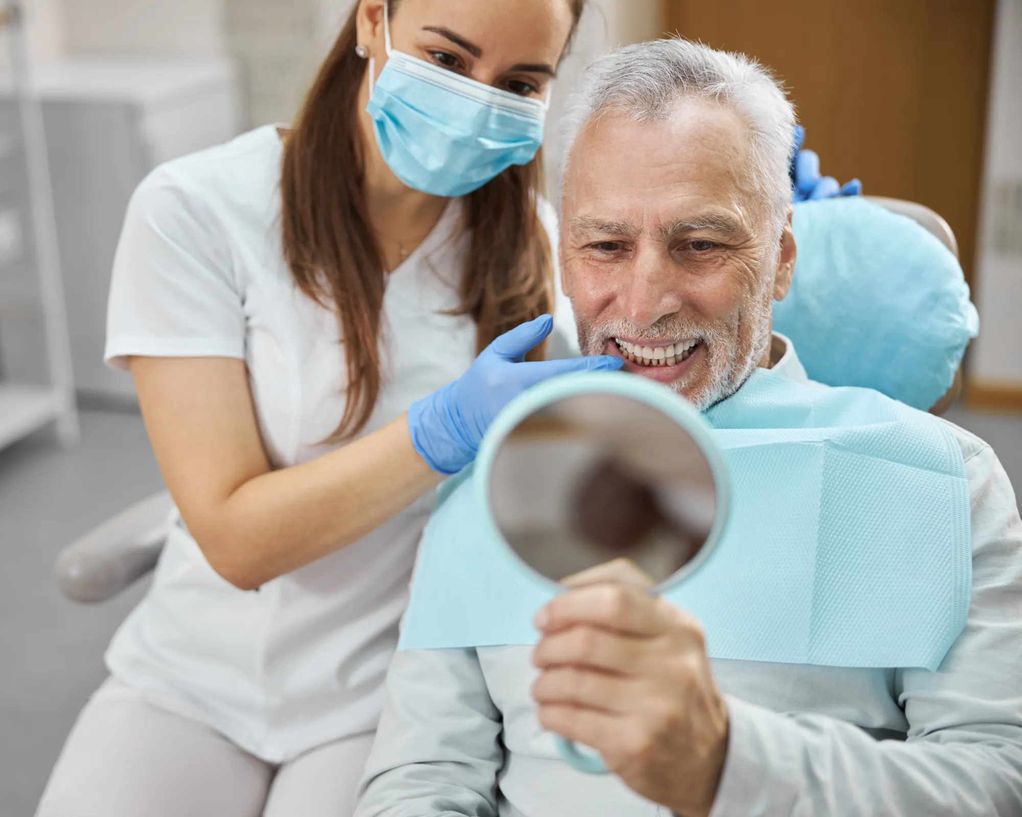 Dentist wearing a mask and gloves showing an older man his teeth as he smiles holding a mirror.