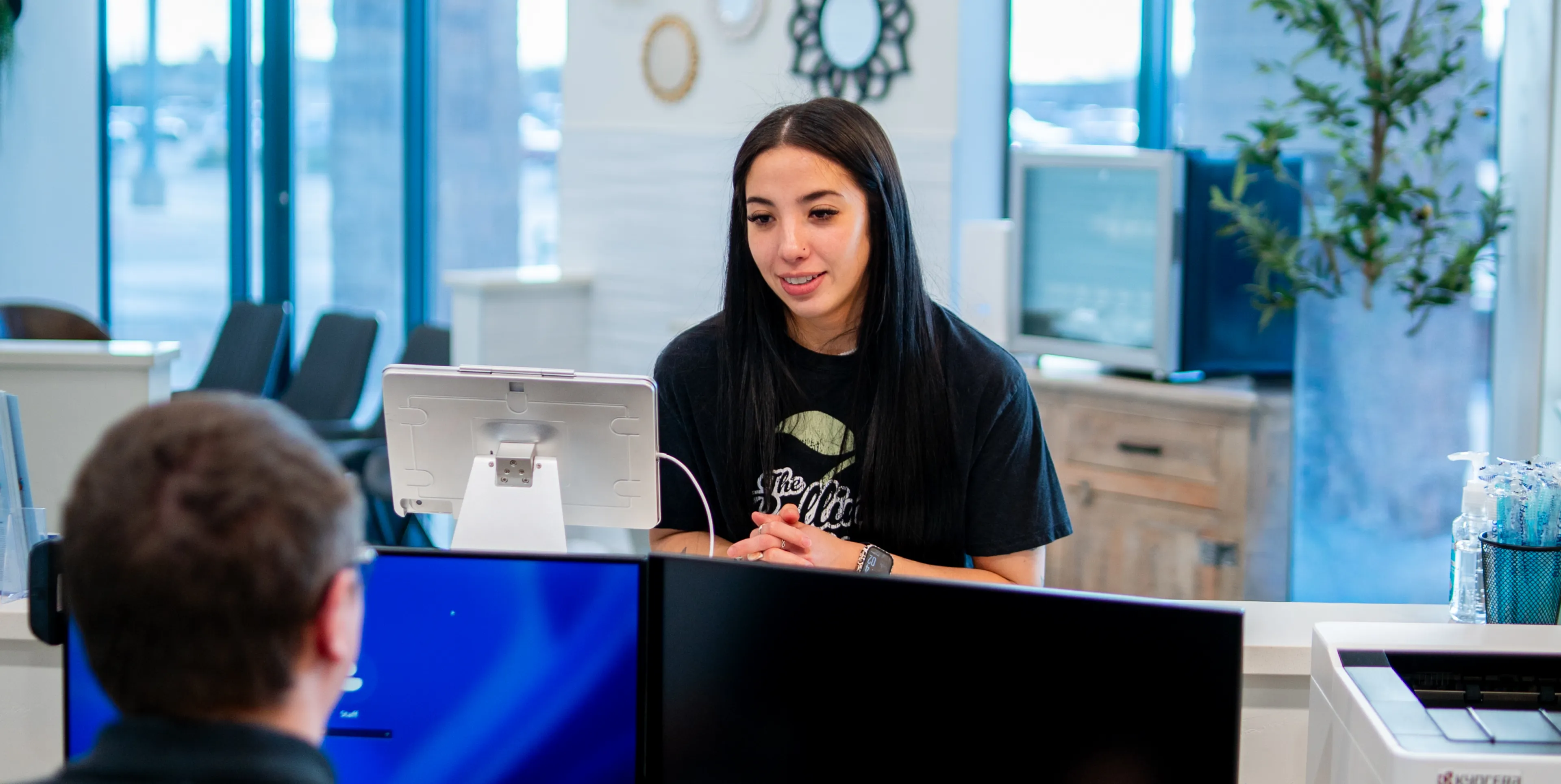 Young woman with long dark hair interacting across a reception desk from a person with short hair in an office setting with computers and large windows in the background.