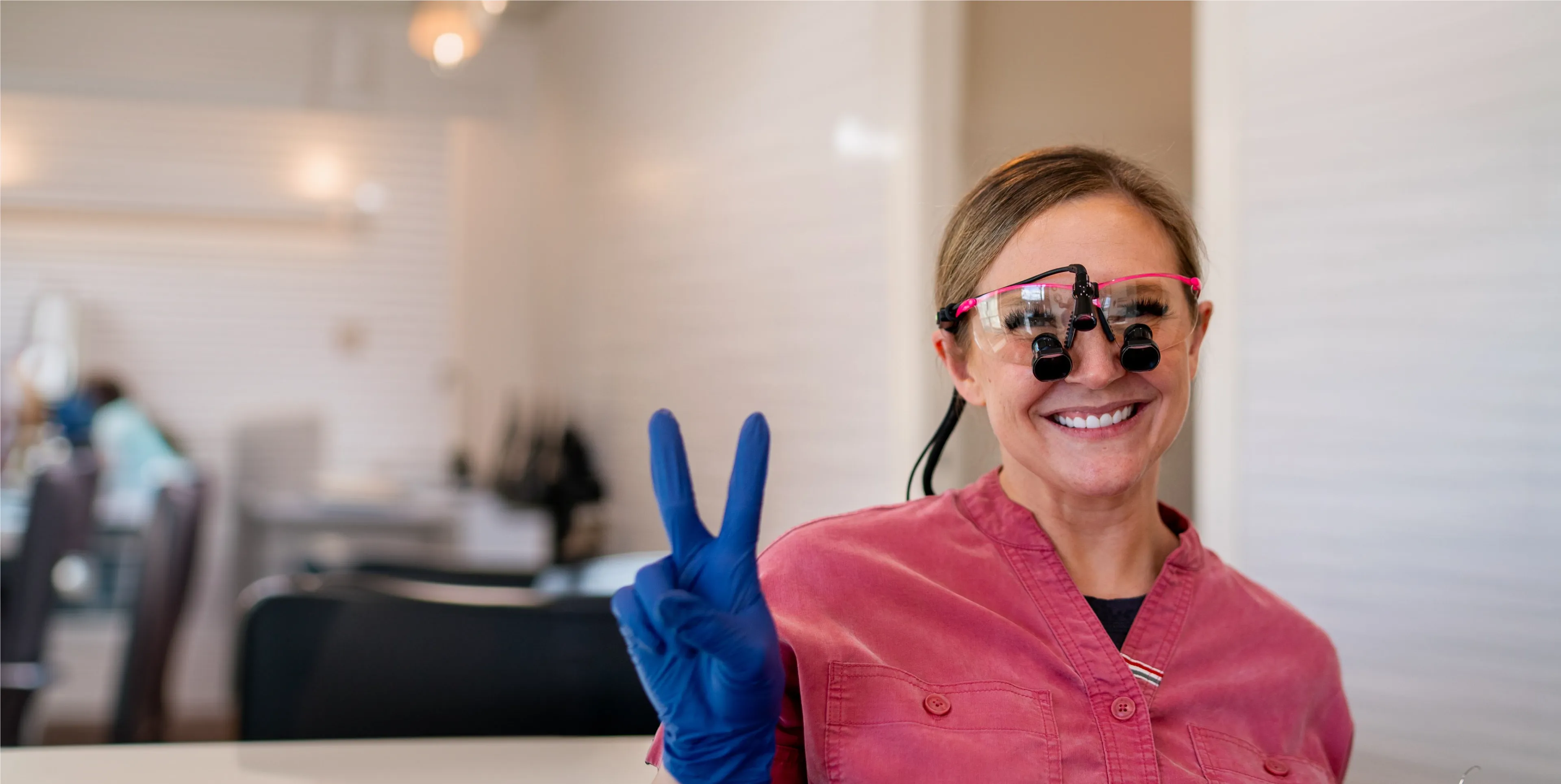 Smiling female dentist wearing magnifying glasses and blue gloves making a peace sign.