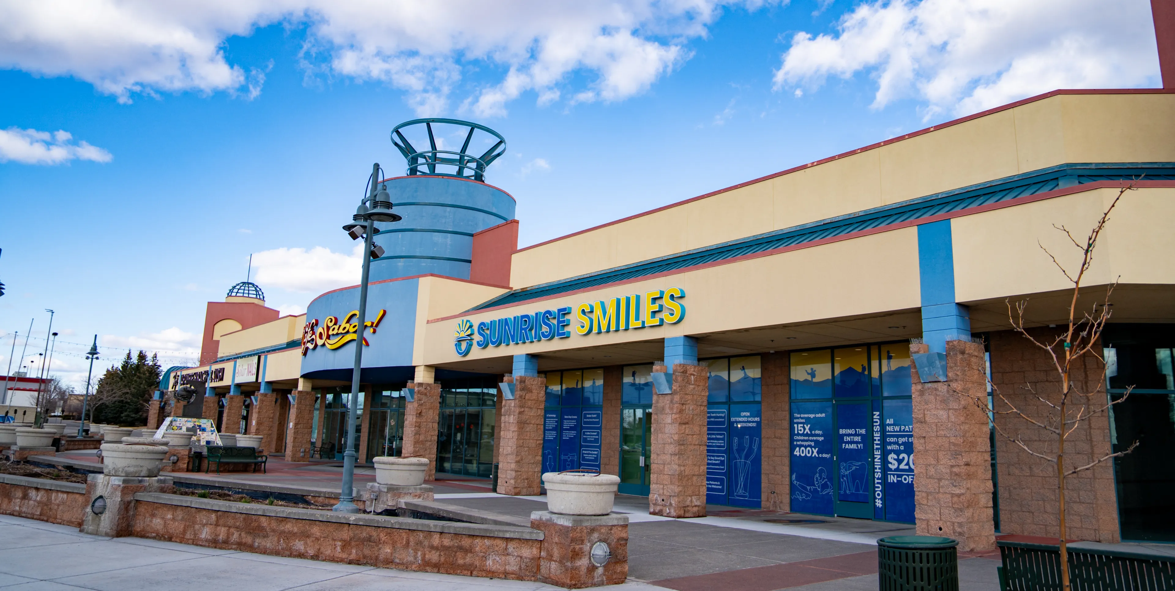 Exterior view of a shopping plaza featuring stores Sunrise Smiles and El Sabor under a partly cloudy blue sky.
