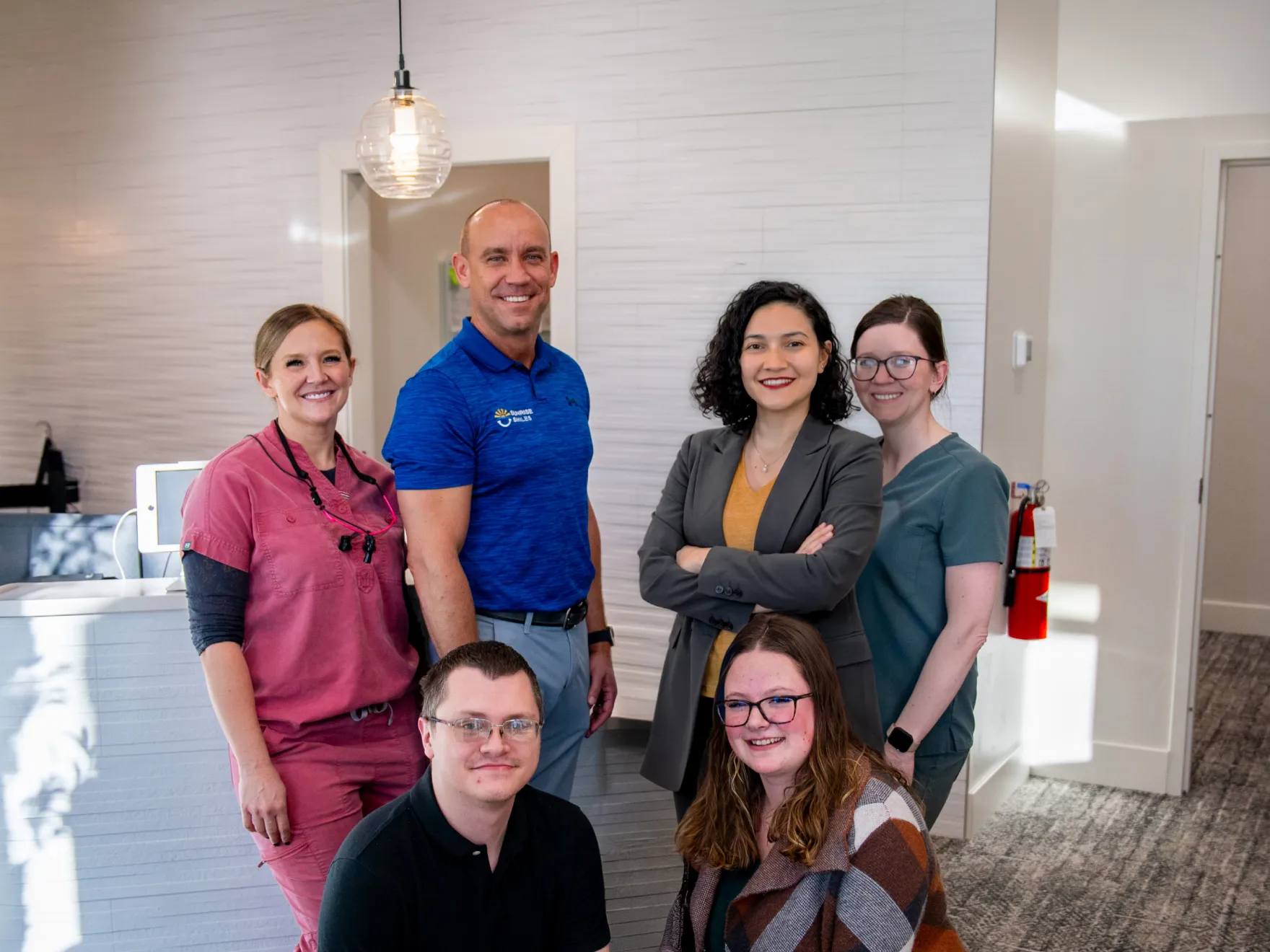 Group of six smiling professionals, including medical staff in scrubs and business attire, standing in a modern office setting.