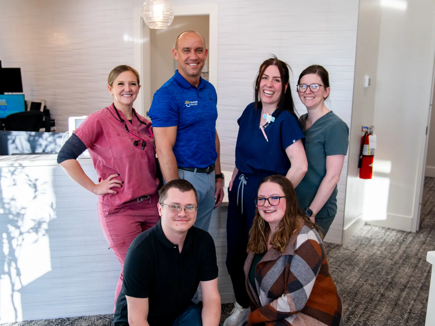 Six smiling dental office staff members posing together inside a modern clinic reception area.